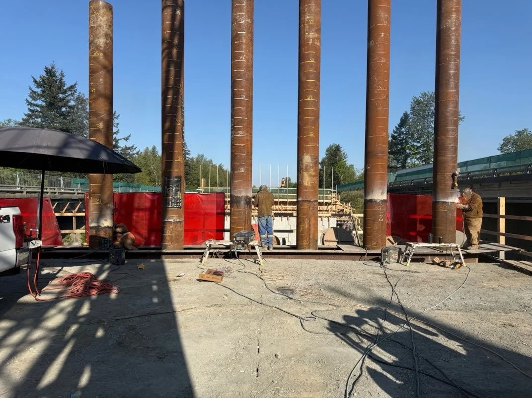 Construction workers installing large rusted steel pipes into a foundation, with red barriers and construction tools around.