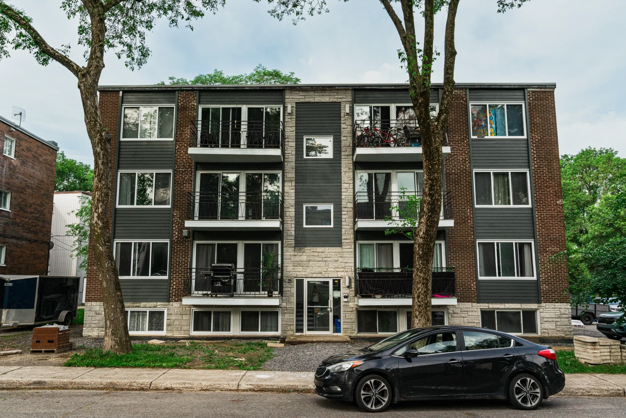 Immeuble résidentiel moderne de quatre étages avec balcons, entouré d'arbres, vue du trottoir et d'une voiture parking devant