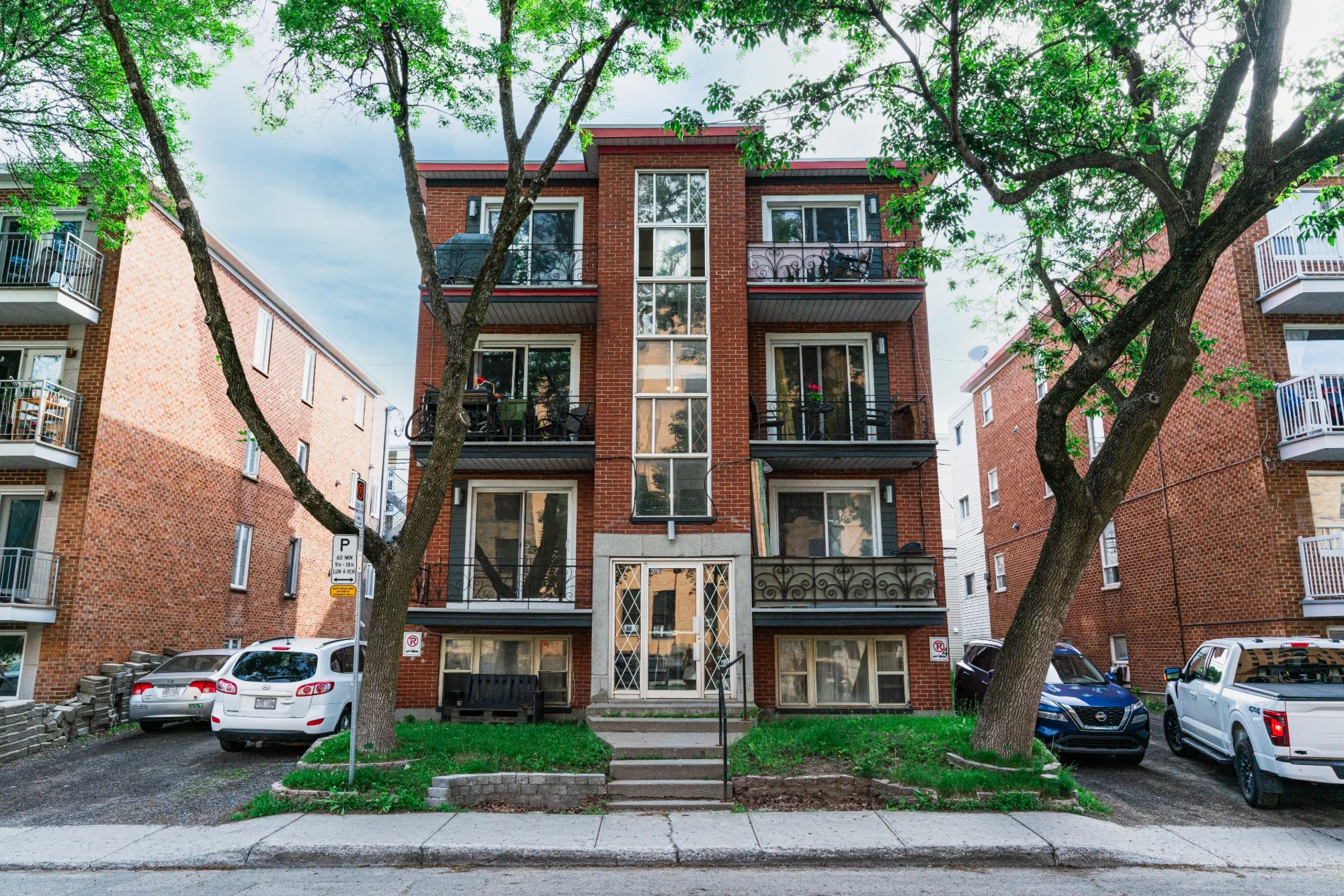 Un immeuble résidentiel en brique rouge avec plusieurs balcons, entouré d'arbres feuillus, avec des voitures garées devant et un panneau de stationnement dans la rue.