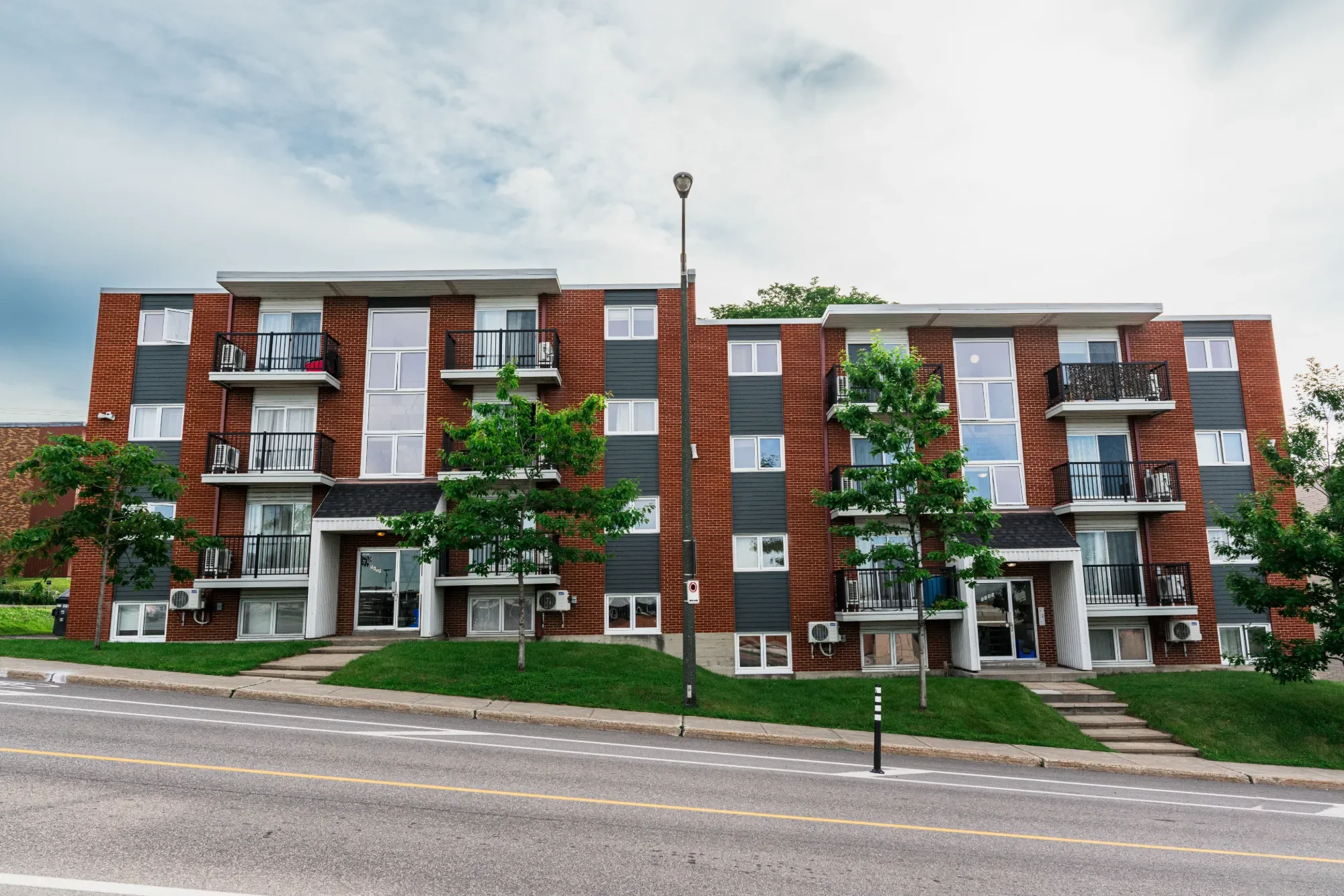 Immeuble résidentiel en briques rouges avec plusieurs balcons, arbres et lampadaire devant, rue en premier plan, ciel partiellement nuageux.