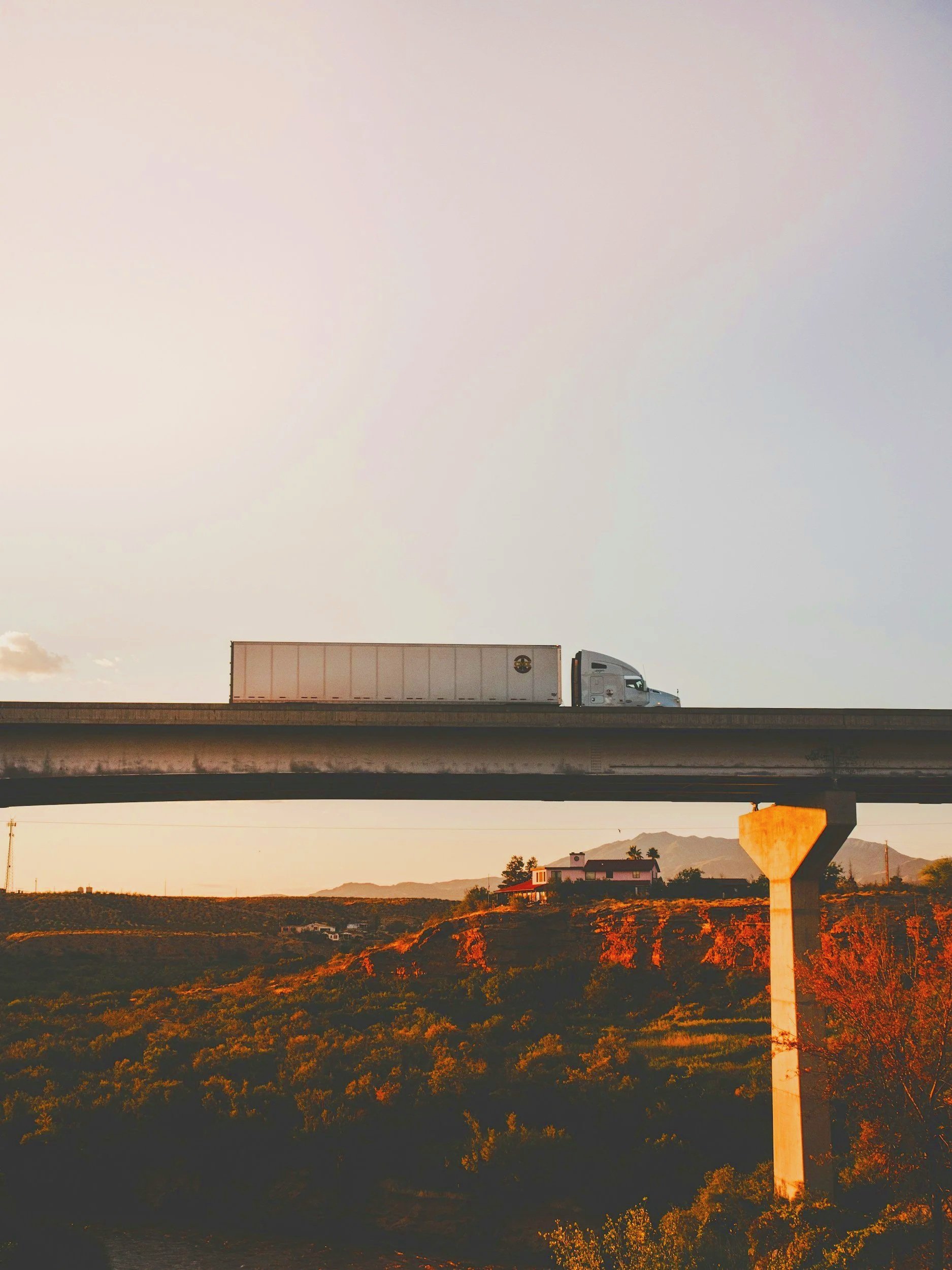 A highway overpass with a semi-truck driving on top during sunset, with a landscape of houses, trees, and mountains in the background.