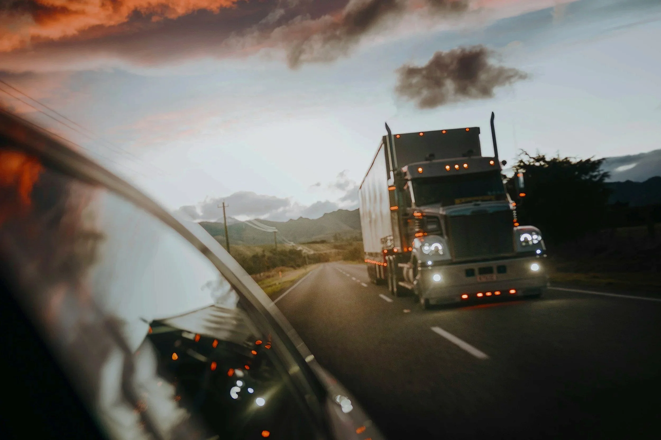 A highway scene with a semi-truck driving on the road during sunset or dusk, with dark smoky clouds in the sky and mountains in the background.