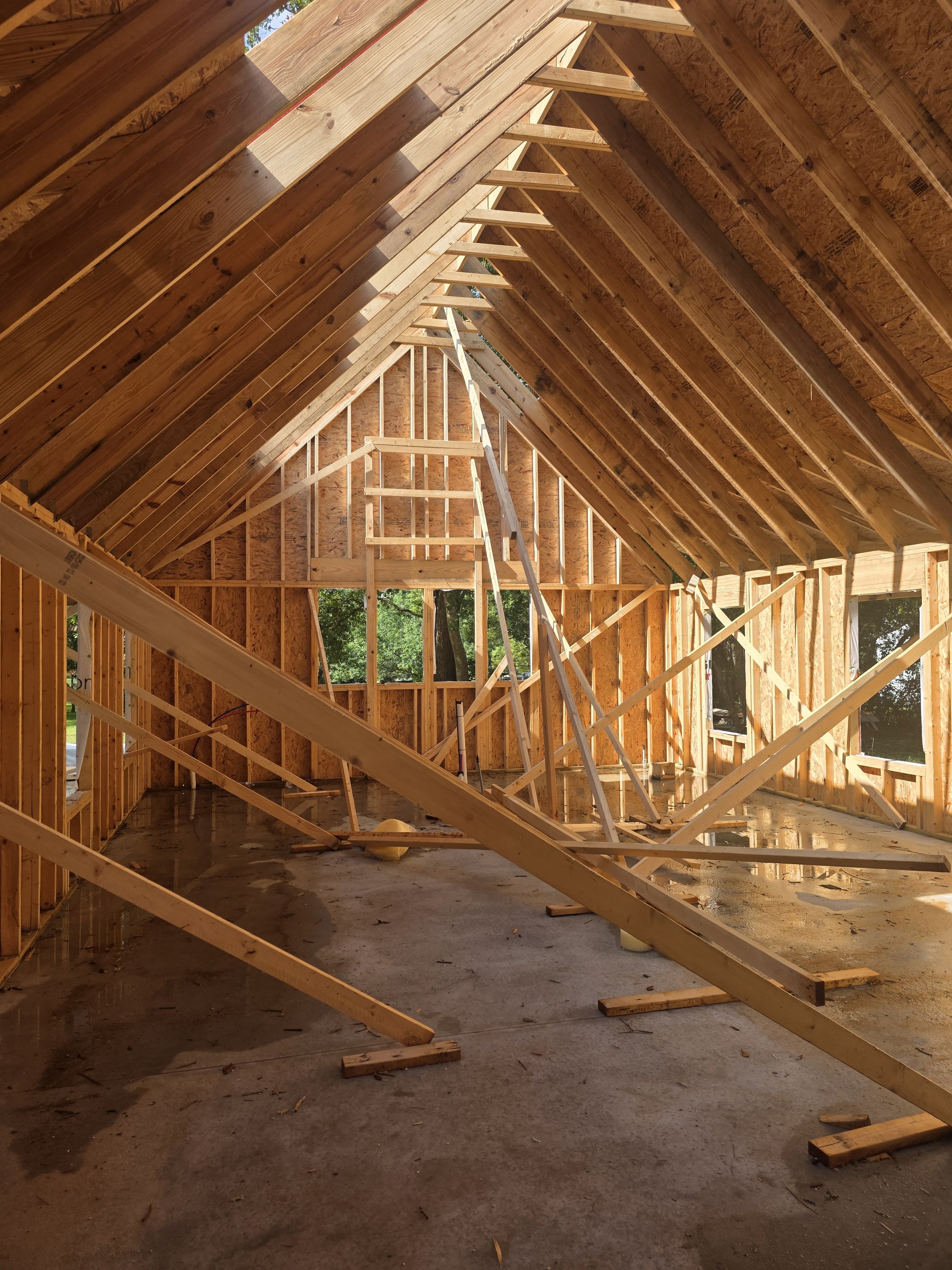 The interior of a house under construction with exposed wooden framing, rafters, and a concrete floor. There are temporary supports and plywood sheathing visible, with sunlight coming through the open roof area.
