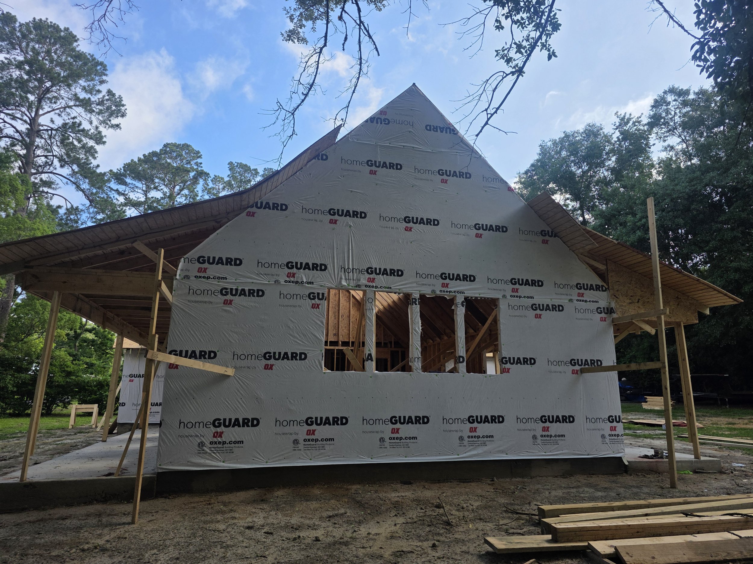 Front view of a house under construction with a sloped roof, covered in 'homeGUARD' house wrap, surrounded by trees and a partly cloudy sky.