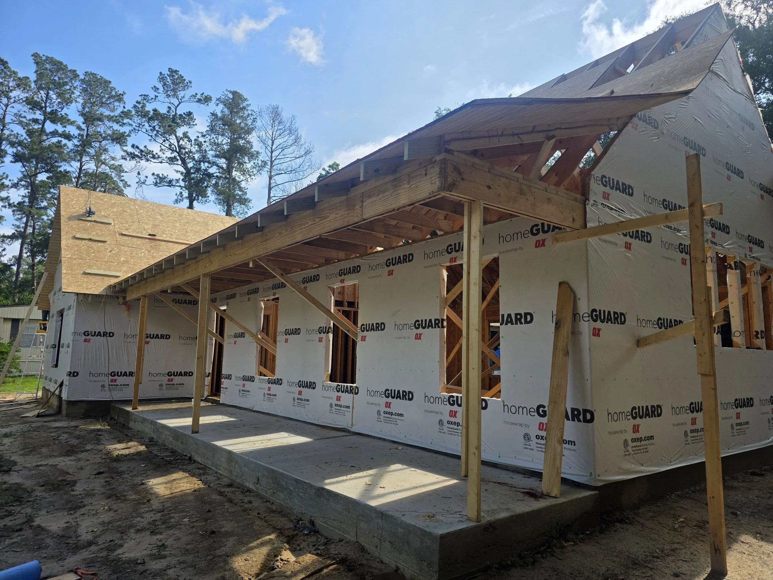 Construction site of a house, showing wooden framework and insulating wrapped walls, with trees and a blue sky in the background.
