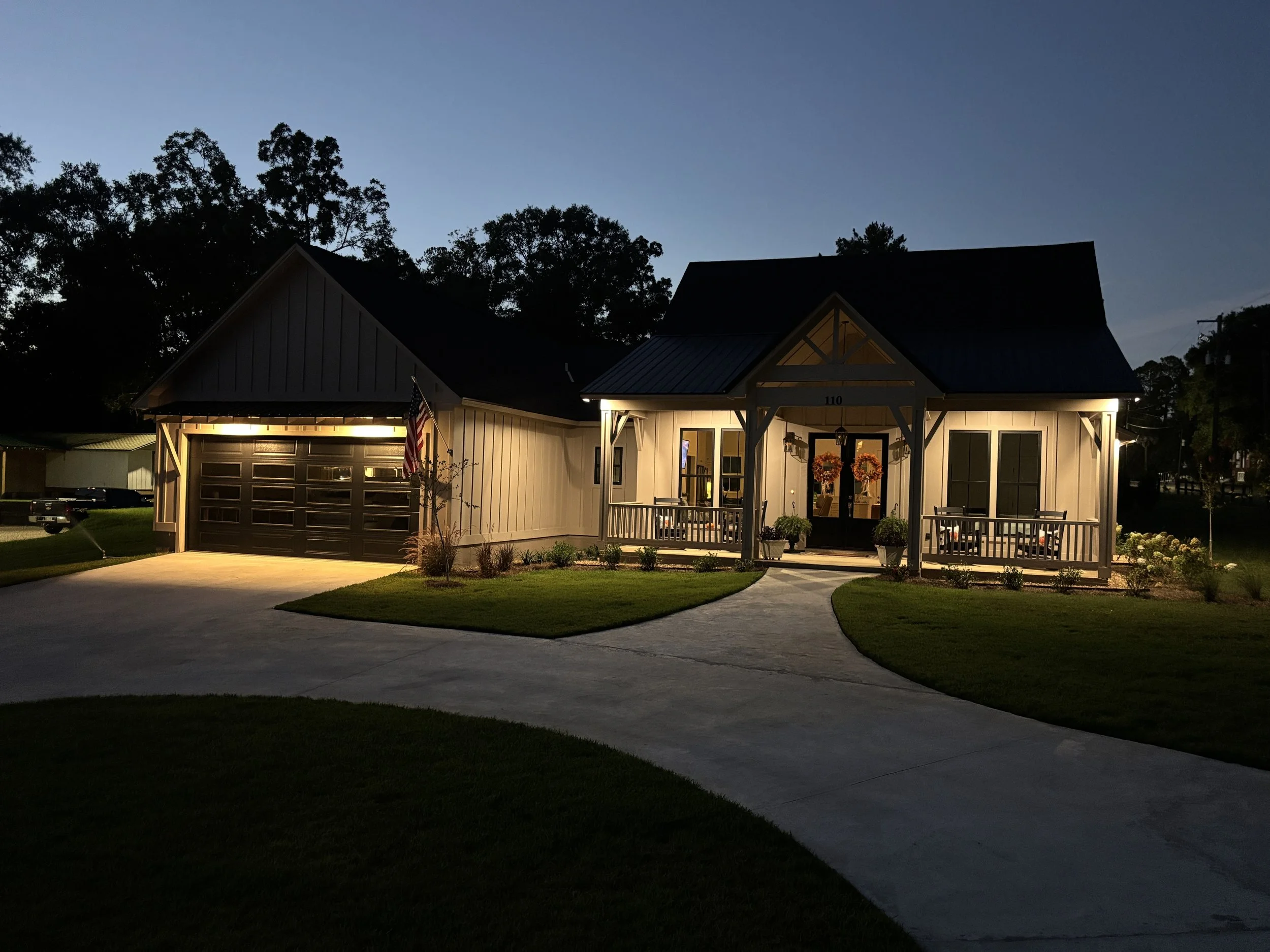 A house at dusk with outdoor lights on, front porch decorated with wreaths, and an American flag. Garage and driveway with lawn and trees in background.