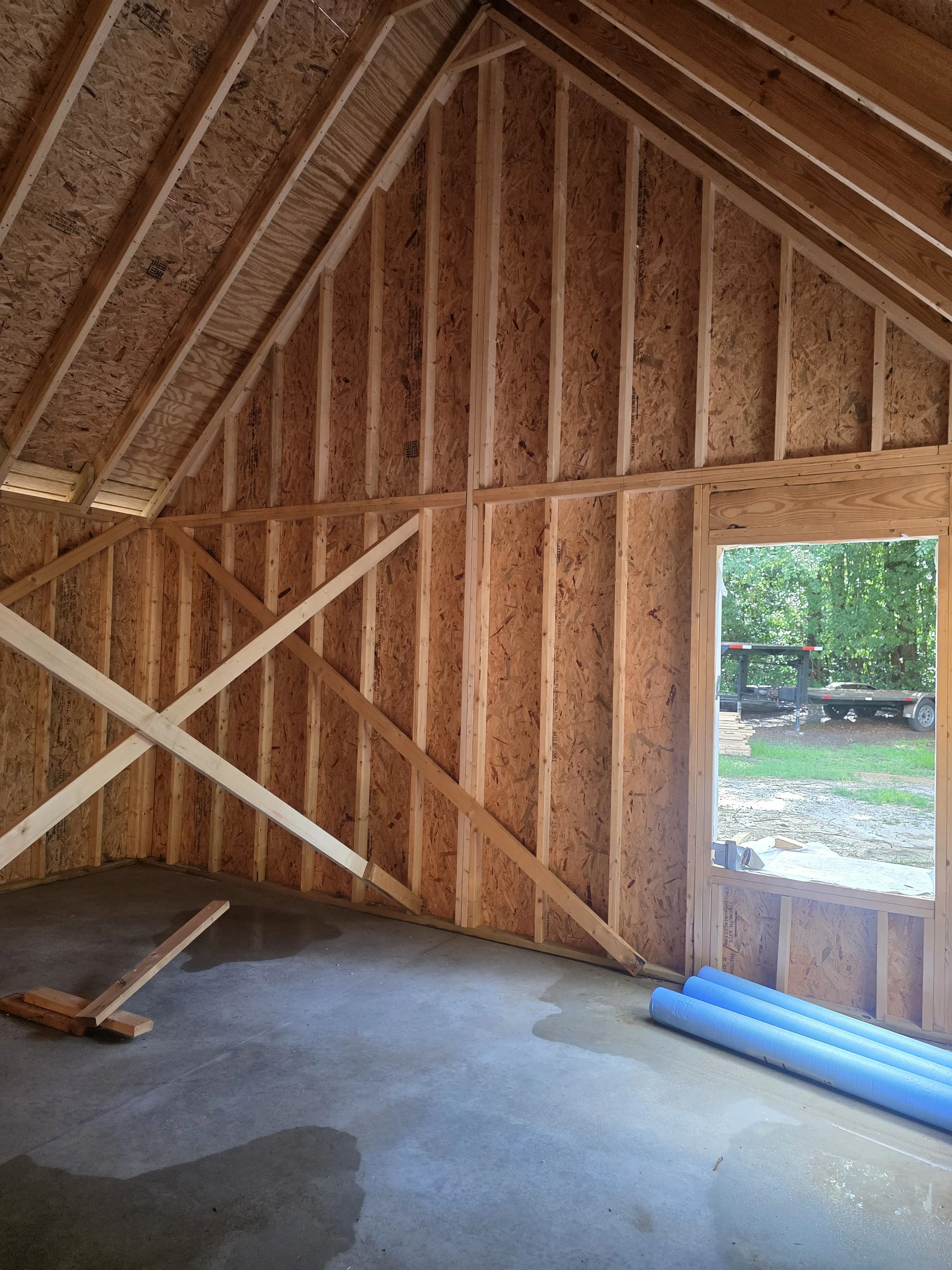 Interior of a building under construction with wooden framing, insulation panels, a concrete floor, and a large open doorway revealing outdoor construction area with trailer and trees.