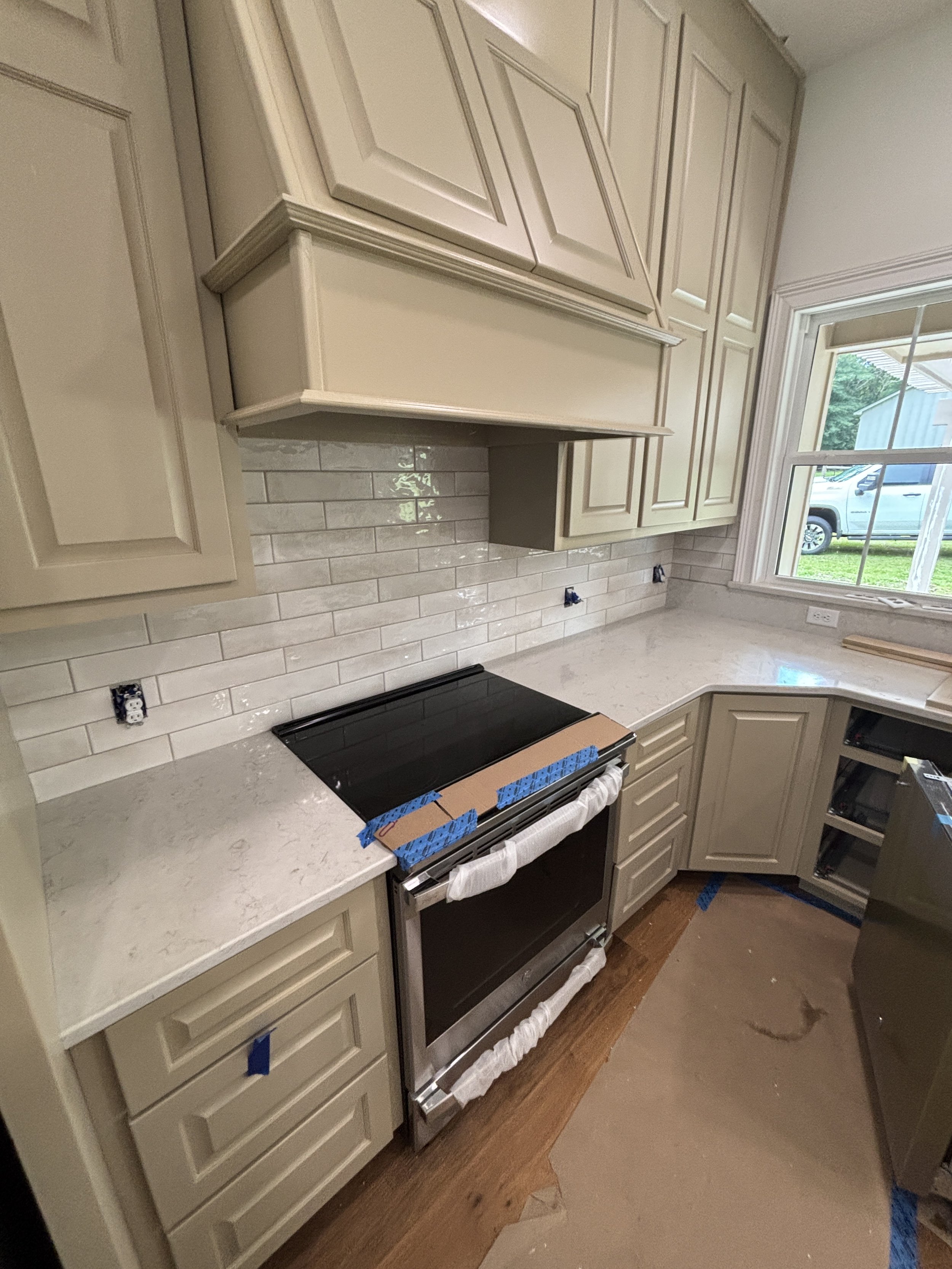 A kitchen with beige cabinets, a granite countertop, and a white subway tile backsplash. There are unfinished electrical outlets and a stovetop under construction, with protective blue tape and packaging materials visible.