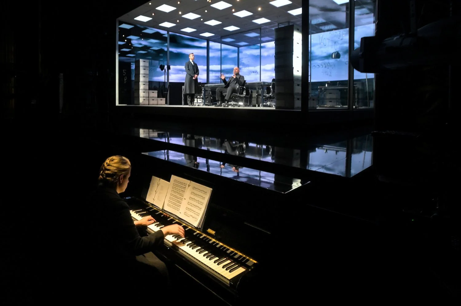 A woman playing piano in a dark room, watching a theatrical performance on a stage through a glass window with a blue sky background.