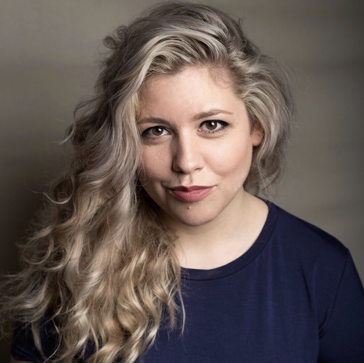 A young woman with wavy blonde hair and brown eyes wearing a navy blue shirt, looking directly at the camera with a slight smile, against a neutral background.