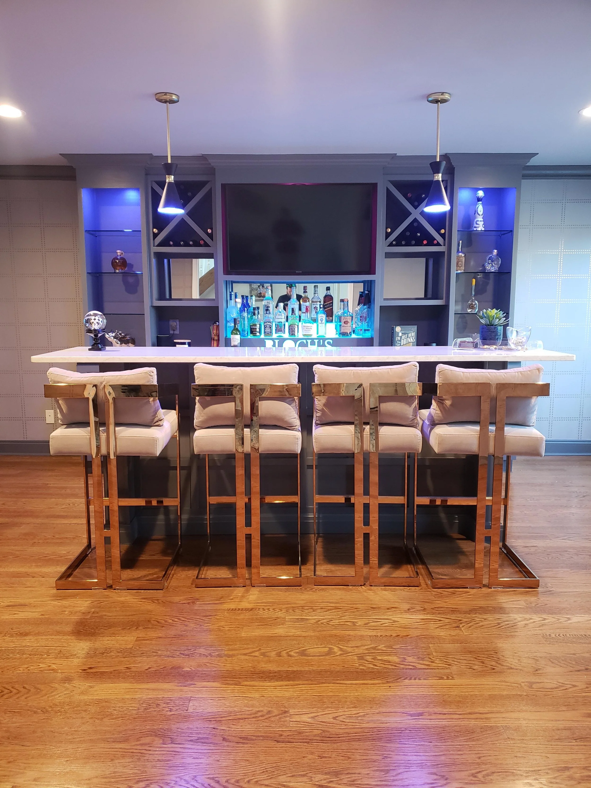 A bar area with a white countertop and high chairs with beige cushions and metallic frames. Behind the bar, there is a built-in shelving unit with bottles of liquor and decorative items liked a small plant and a skull figurine. Two pendant lights hang from the ceiling illuminating the space, with a large flat-screen television mounted in the center of the shelving unit.