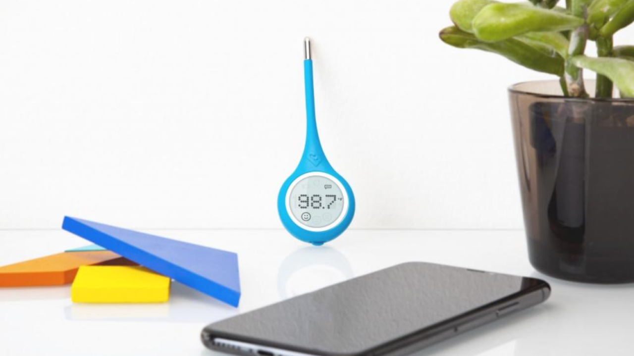 A white surface holding a blue digital thermometer displaying 98.7°F, a gray smartphone, multicolored sticky notes, and a potted plant with green leaves in a black pot.