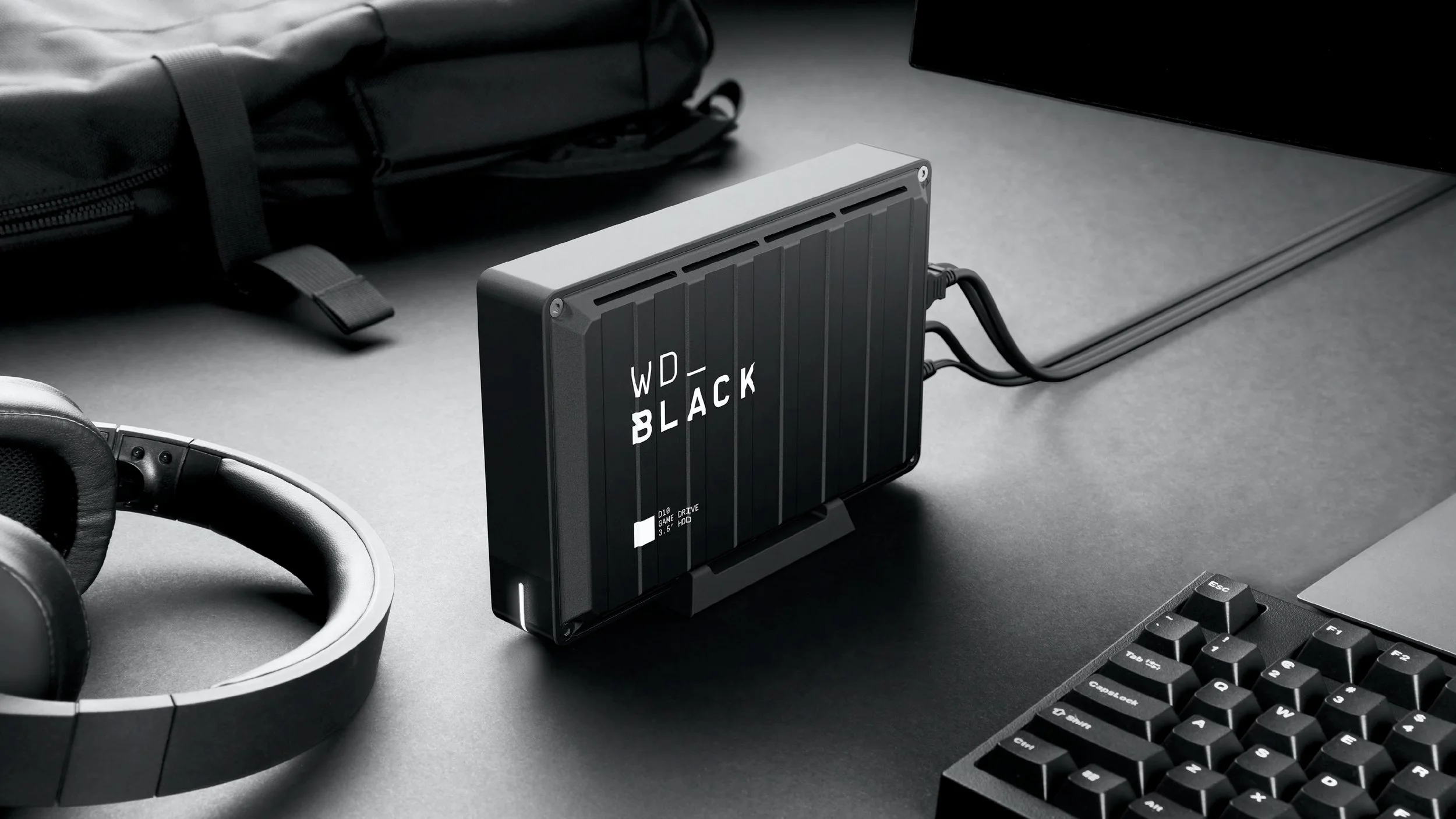 A black external hard drive labeled 'WD Black' connected with cables on a dark desk, alongside a black keyboard, headphones, and a backpack.