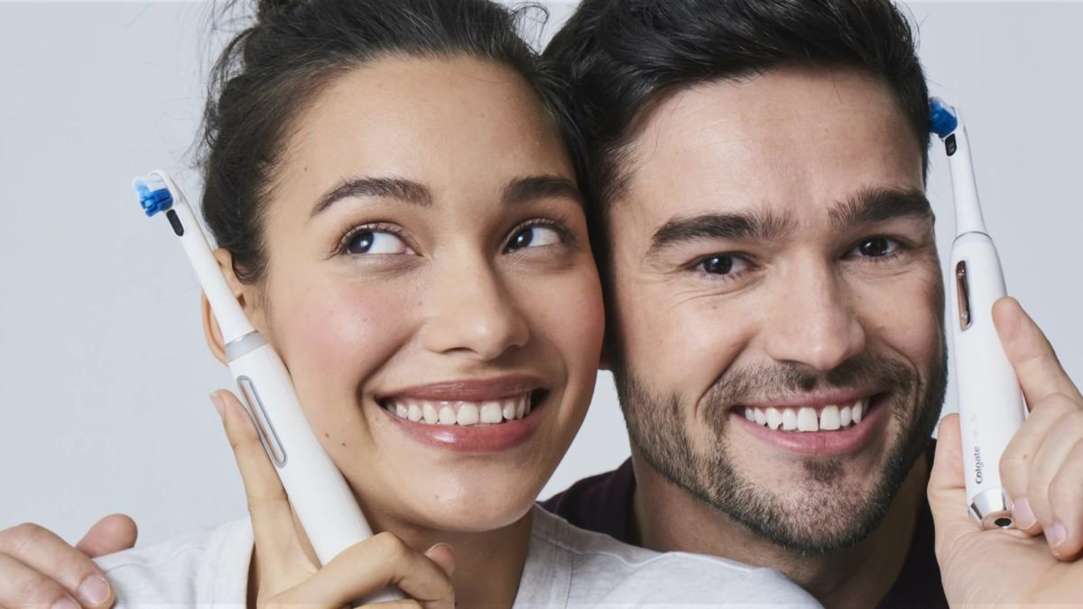 A smiling woman and man holding electric toothbrushes near their faces, standing closely together against a plain background.