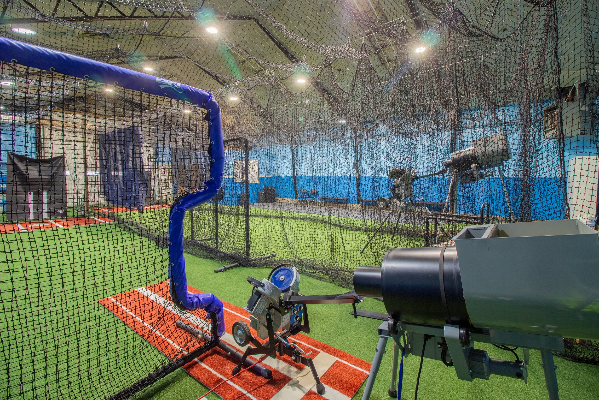 A batting cage setup inside a sports training facility. The cage has netting, batting tee, a pitching machine, and various sports equipment, with artificial turf on the floor and bright overhead lighting.