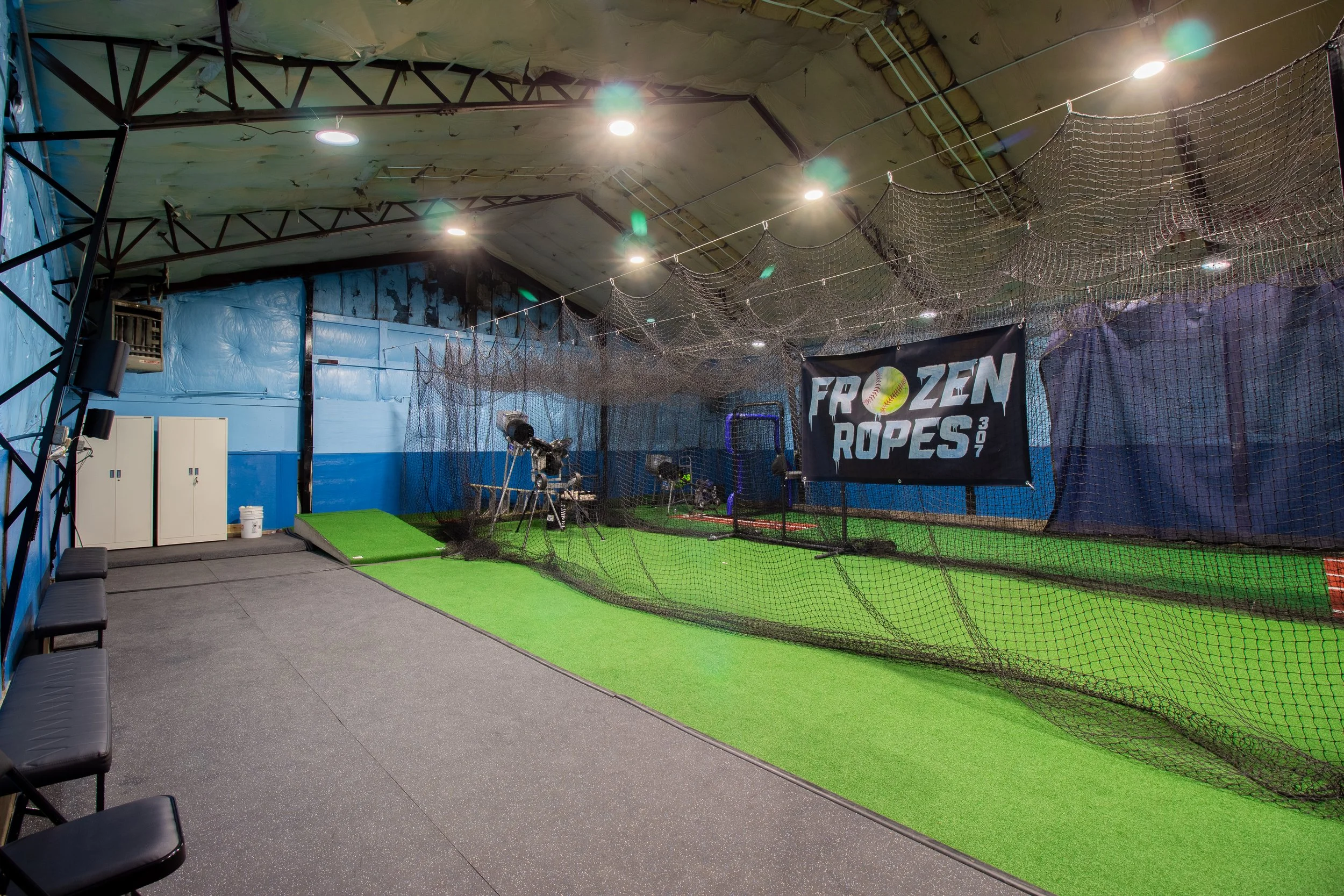Indoor batting cage with green turf, black nets, and a blue background banner reading 'Frozen Ropes' with a baseball graphic, several baseballs on the ground, chairs on the side, and exercise equipment in the background.