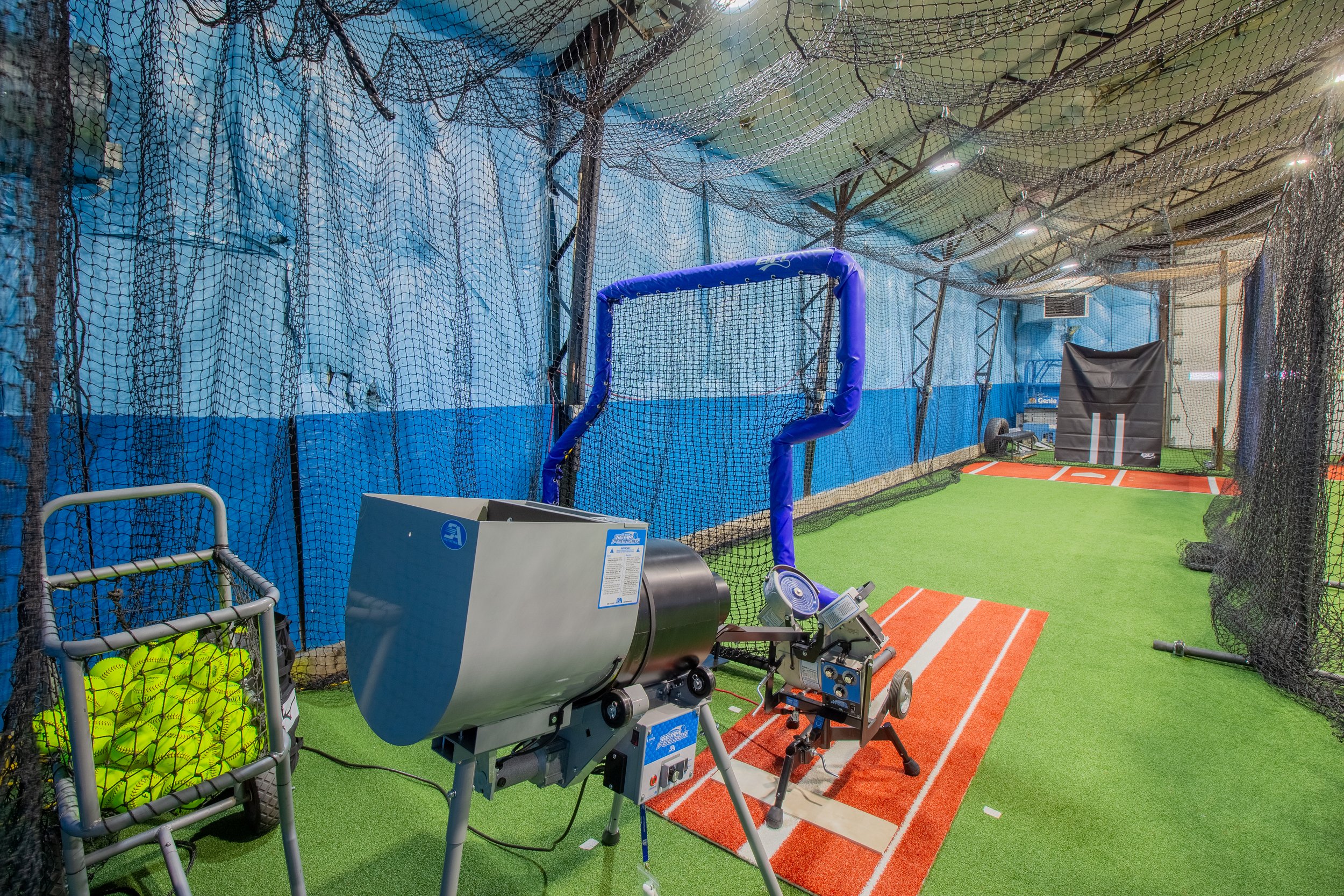 Indoor baseball or softball training facility with netting, artificial turf, and pitching machines, including a pitching mound, tennis balls in a basket, and sports equipment.