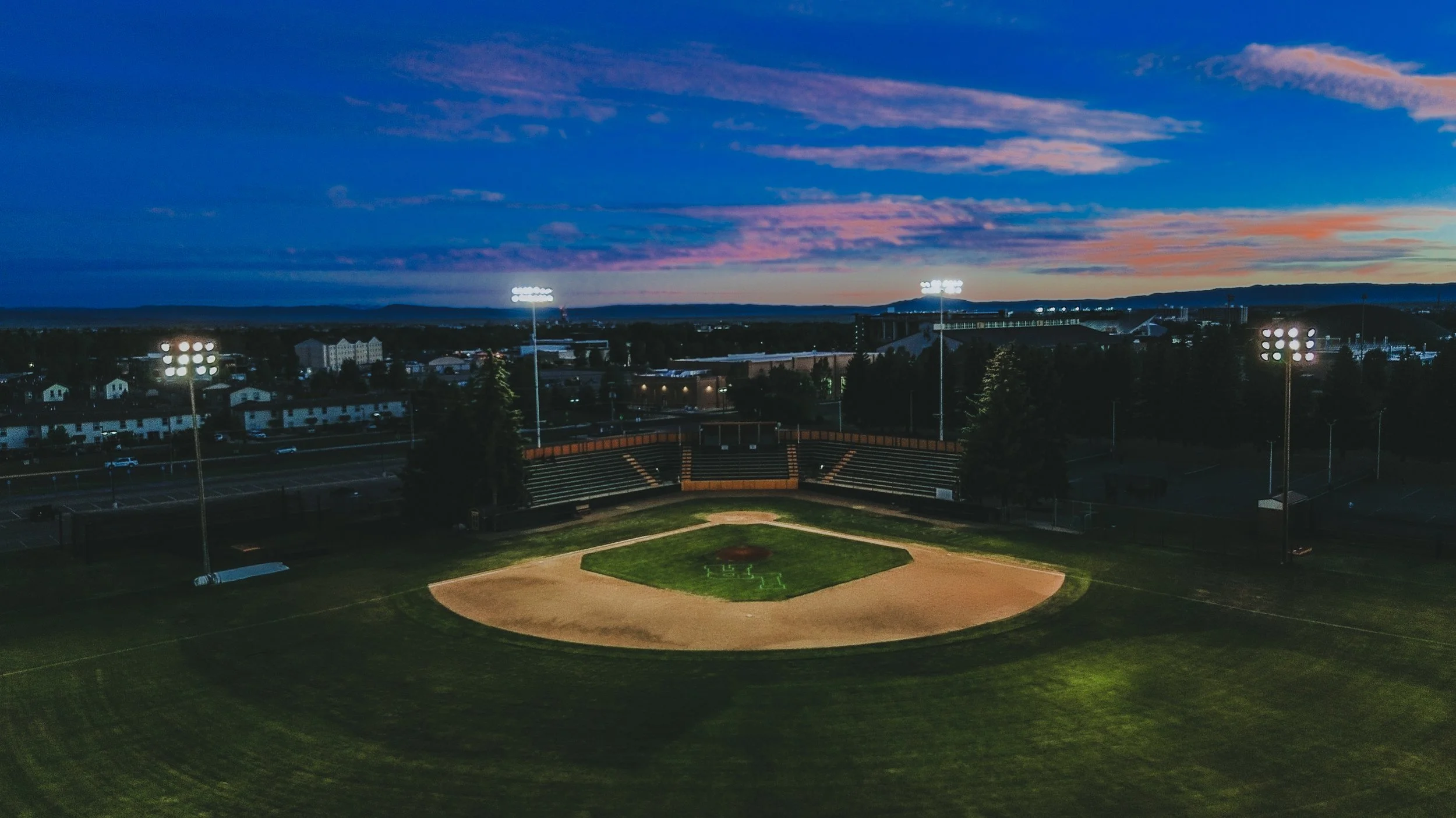 View of a baseball field at dusk with stadium lights on, a hill with buildings in the background, and a colorful sky with pink, purple, and blue clouds.
