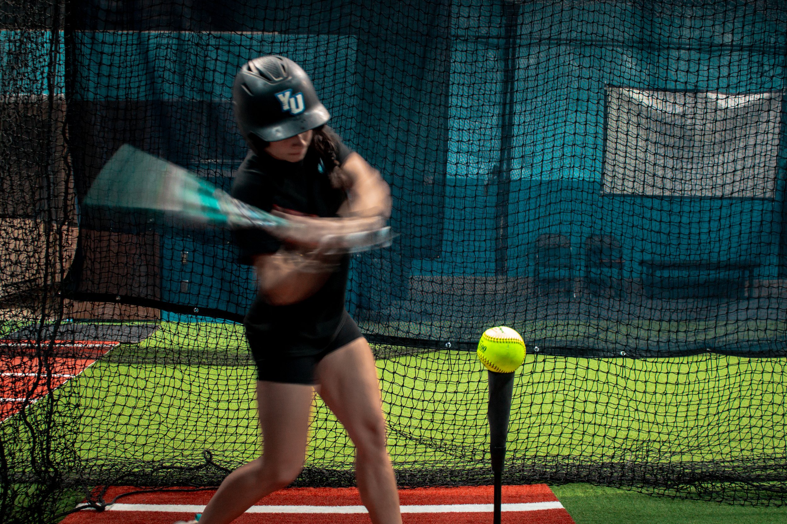 A female softball batter practicing inside a batting cage, wearing a black helmet with a white logo and black shirt, with a yellow softball on a tee.