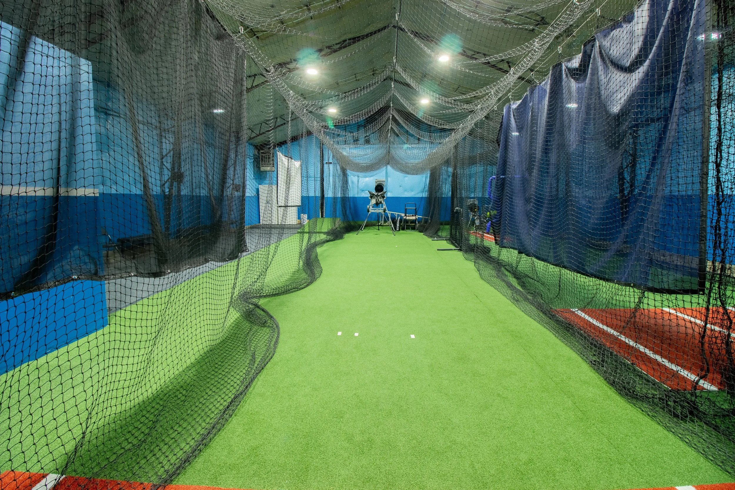 Indoor batting cage with black netting and green flooring, equipped with pitching machines and batting tees.