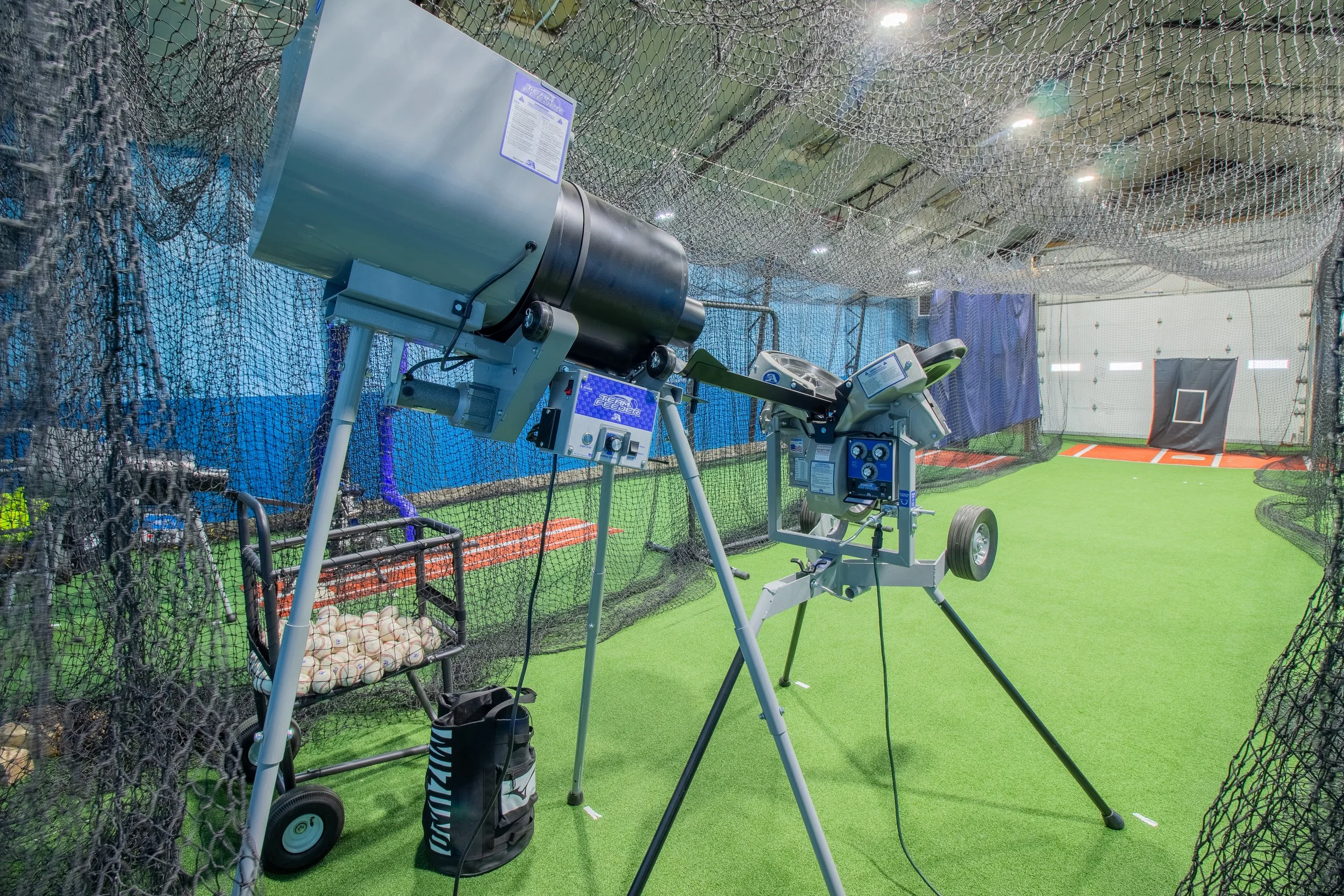 Indoor baseball or softball training area with netting, green artificial turf, and batting equipment including a pitching machine, ball basket, and a hitting target in the background.