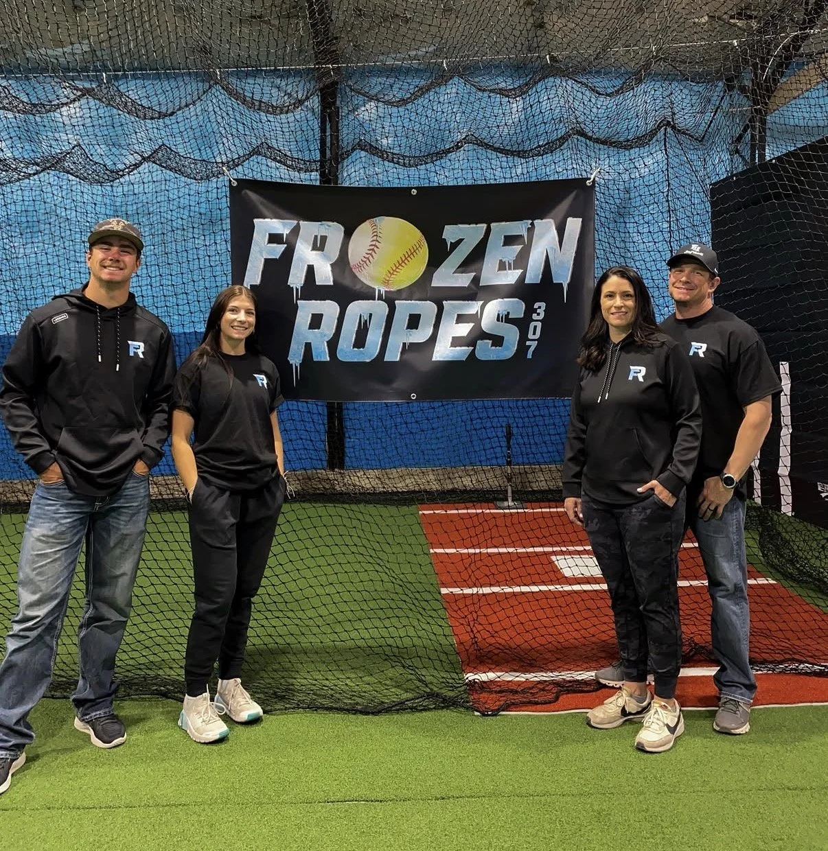 Four people standing inside an indoor baseball training facility in front of a banner that reads 'Frozen Ropes 307'. They are smiling and wearing black hoodies or t-shirts with the letter 'R' on them. The background has a net and a red and white batting cage.