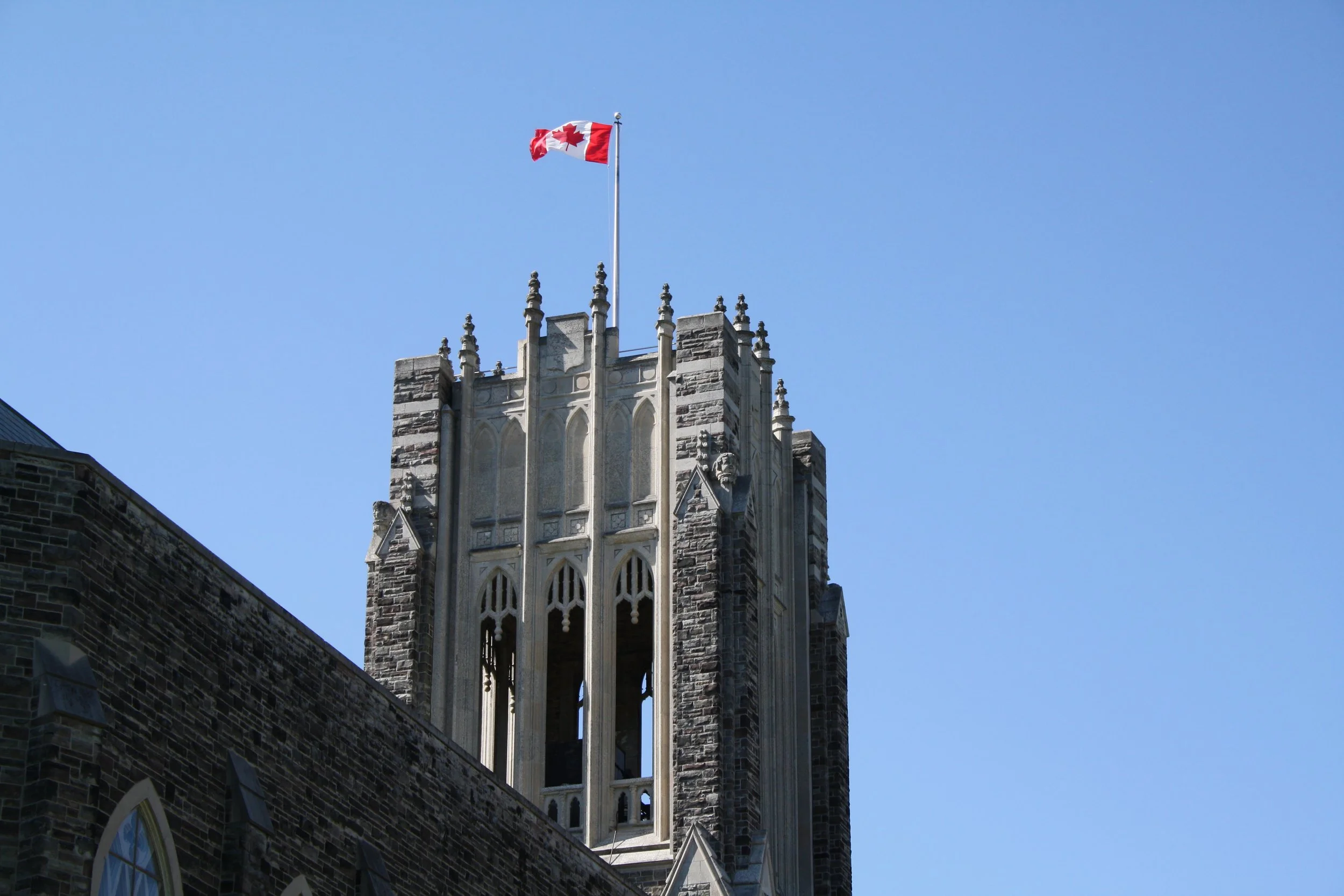 A close-up of a historic stone building with gothic architectural details and a flagpole with the Canadian flag flying, set against a clear blue sky.