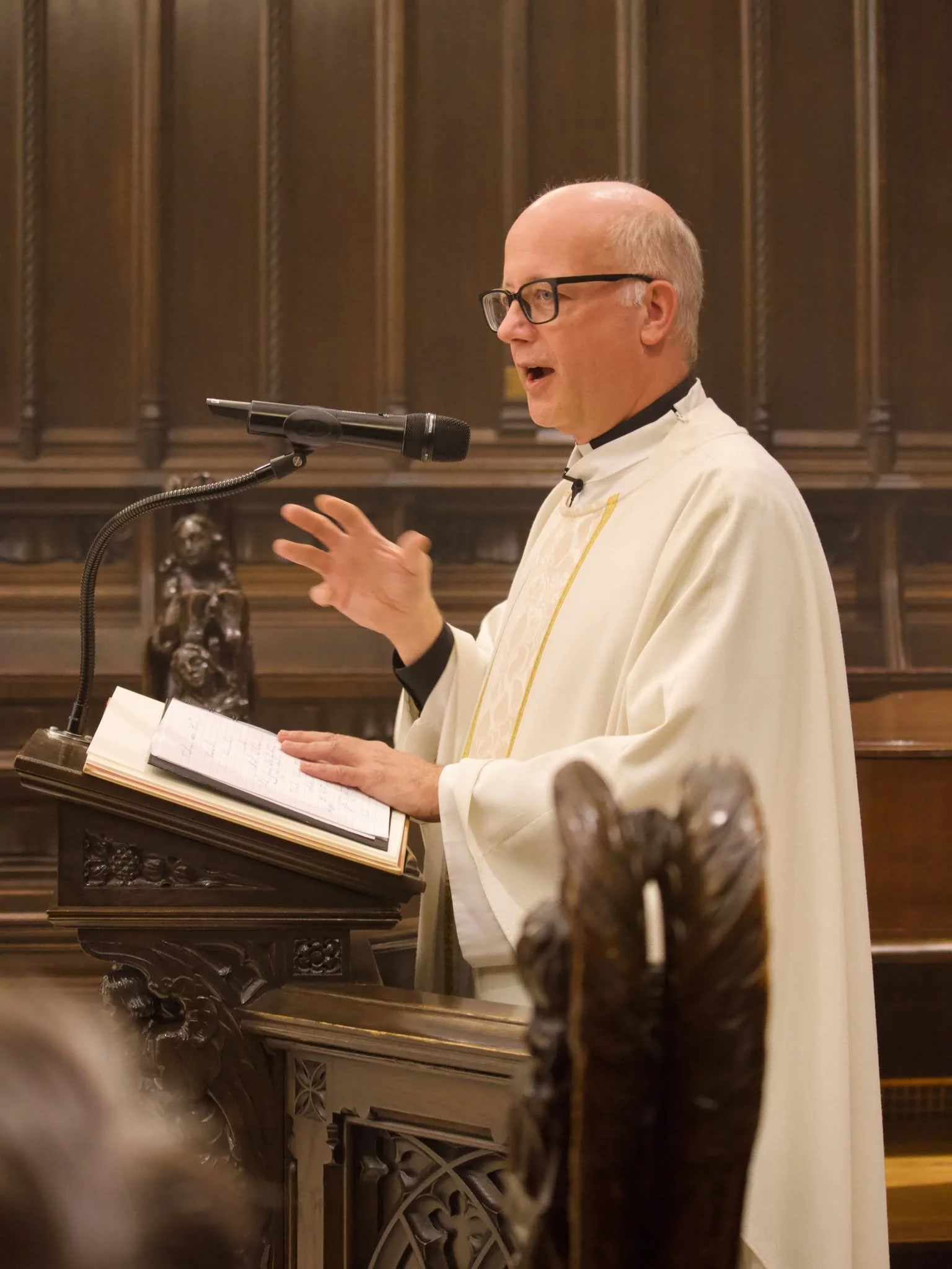 A priest in white robe and glasses speaking into a microphone at a podium with an open book in front of him, standing in a wooden church or cathedral.