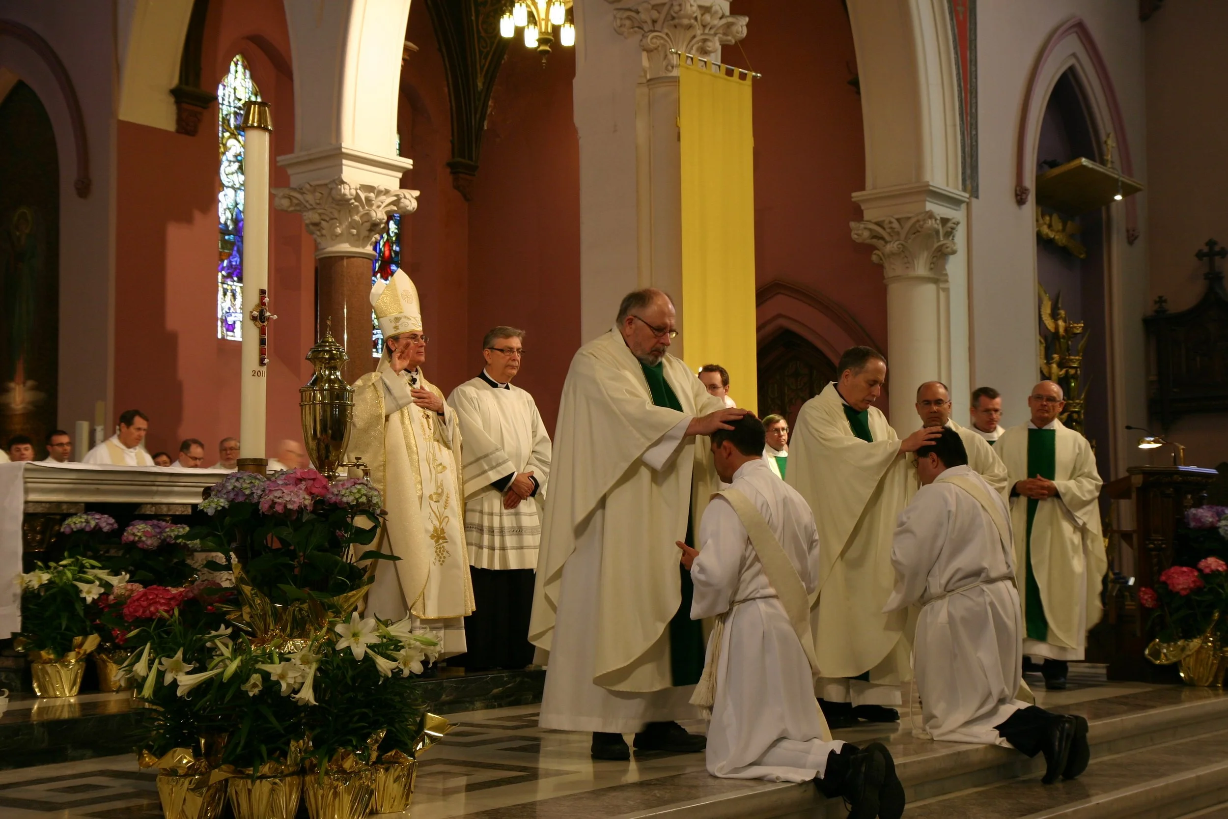 Inside a church, priests and altar servers participate in a religious ceremony. One priest is kneeling, while another priest is laying hands on a kneeling man. Flowers and religious symbols decorate the altar.