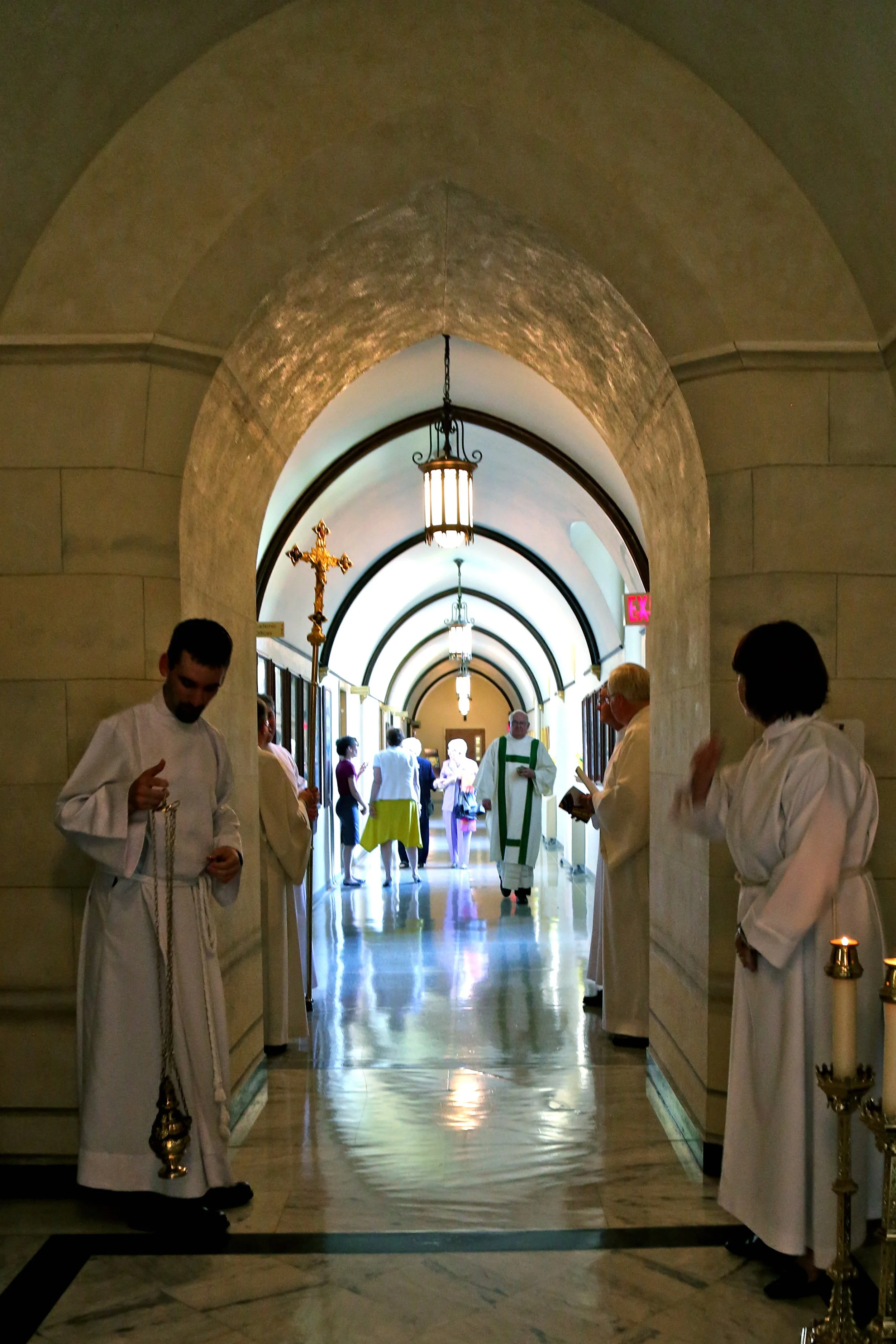Inside a church, priests and congregants gather in a corridor with arched ceilings, decorating with candles and religious symbols.
