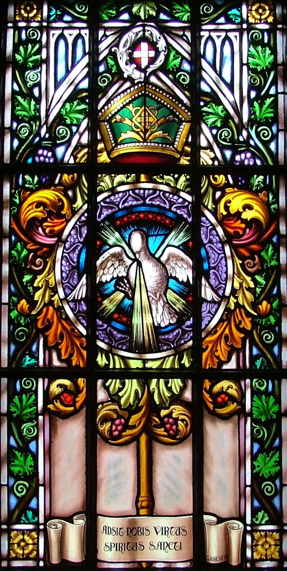 Colorful stained glass window depicting a dove with rays of light, surrounded by ornate floral and leaf patterns, with a scroll at the bottom bearing Latin text.