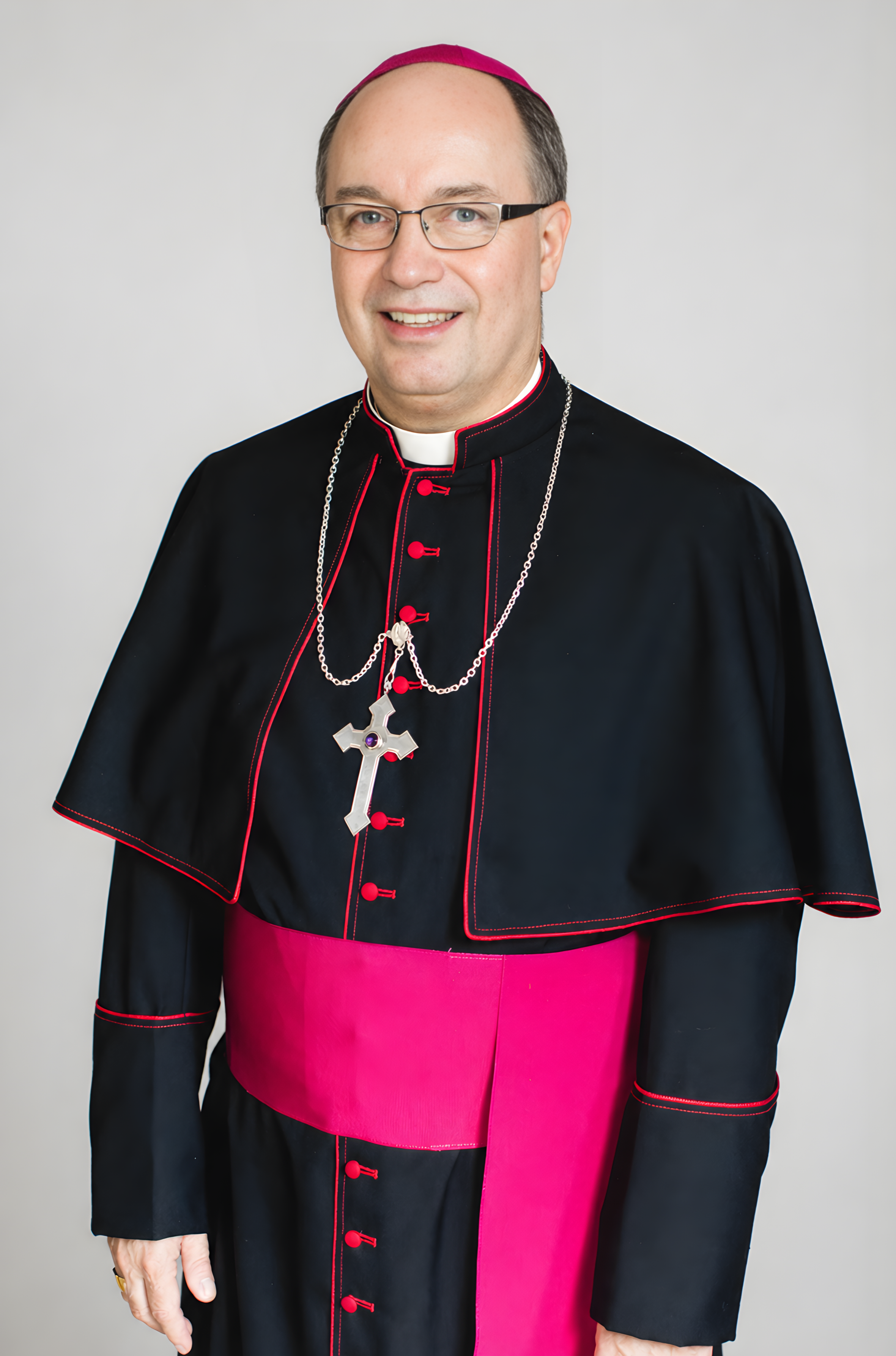 A bishop dressed in traditional clerical attire with a clerical collar, black cassock with red piping and buttons, a pink sash around the waist, a pink zucchetto, and a silver cross necklace, smiling at the camera.