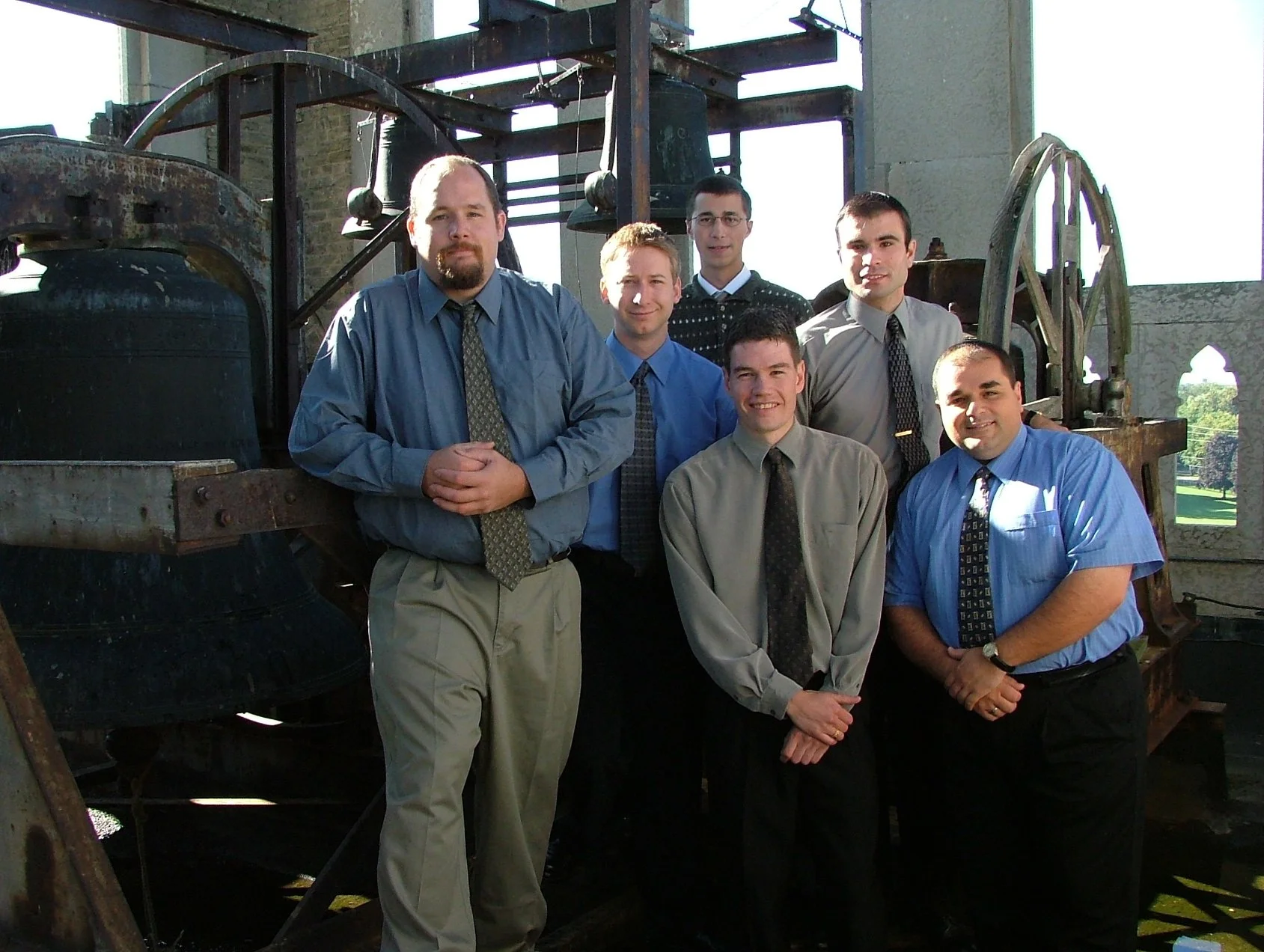 A group of seven men in business casual attire standing in front of a large old bell and machinery inside a church or historic building with stone walls and arched windows.