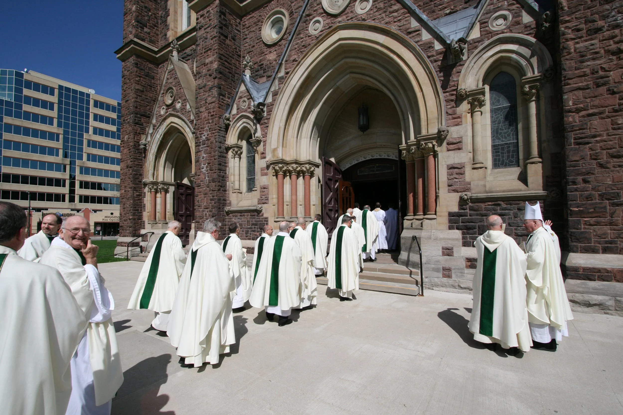 A group of clergy members dressed in white coats with green stoles walking towards the entrance of a stone church with arched doors and stained-glass windows. Several people are smiling or greeting each other on the sidewalk.
