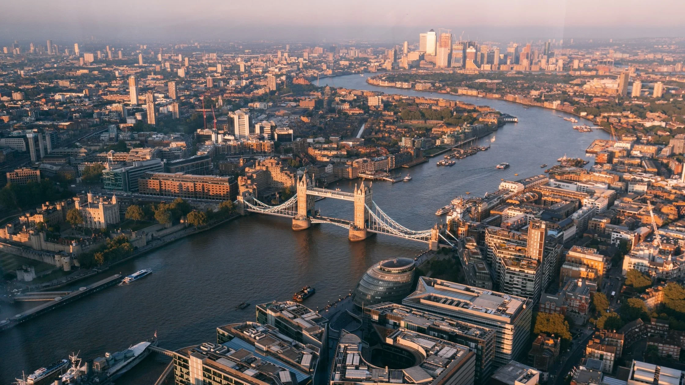 Aerial view of London cityscape at sunset, featuring the Tower Bridge over the River Thames and various high-rise buildings and historic architecture.