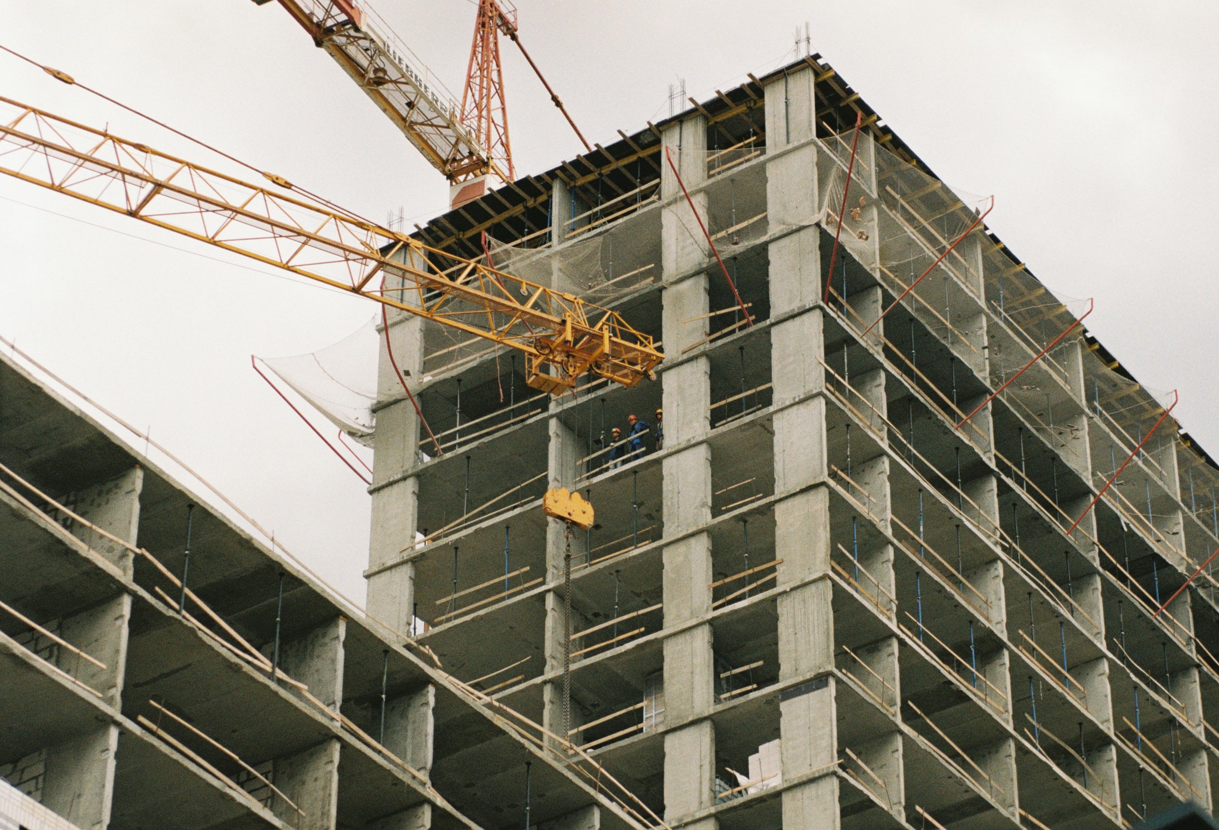 Construction site with a partially built multi-story concrete structure, workers in safety gear, and a yellow tower crane lifting materials.