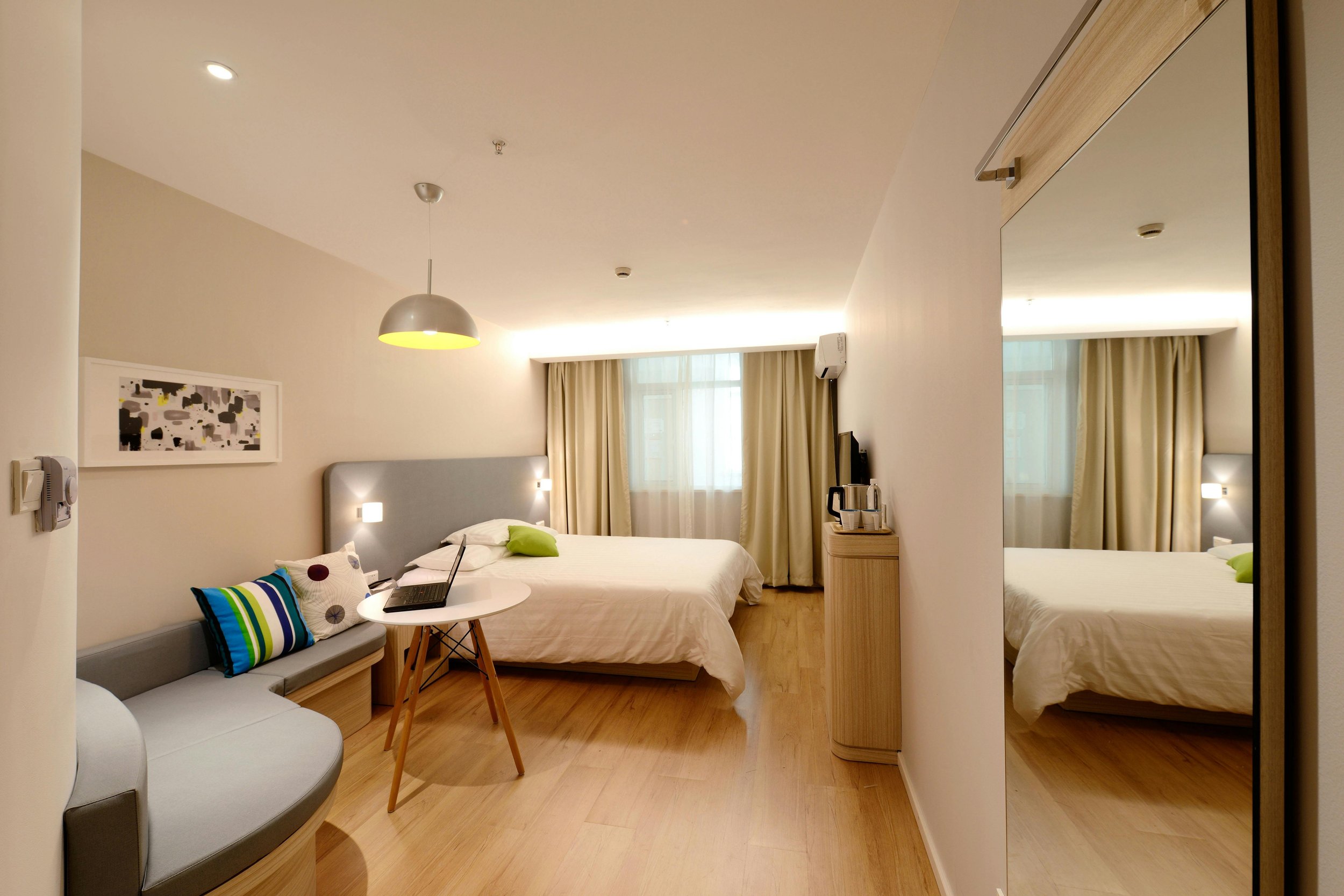 Interior view of a modern hotel room with two beds, a small couch with colorful pillows, a wall mirror, a desk with a laptop, and a window with curtains.
