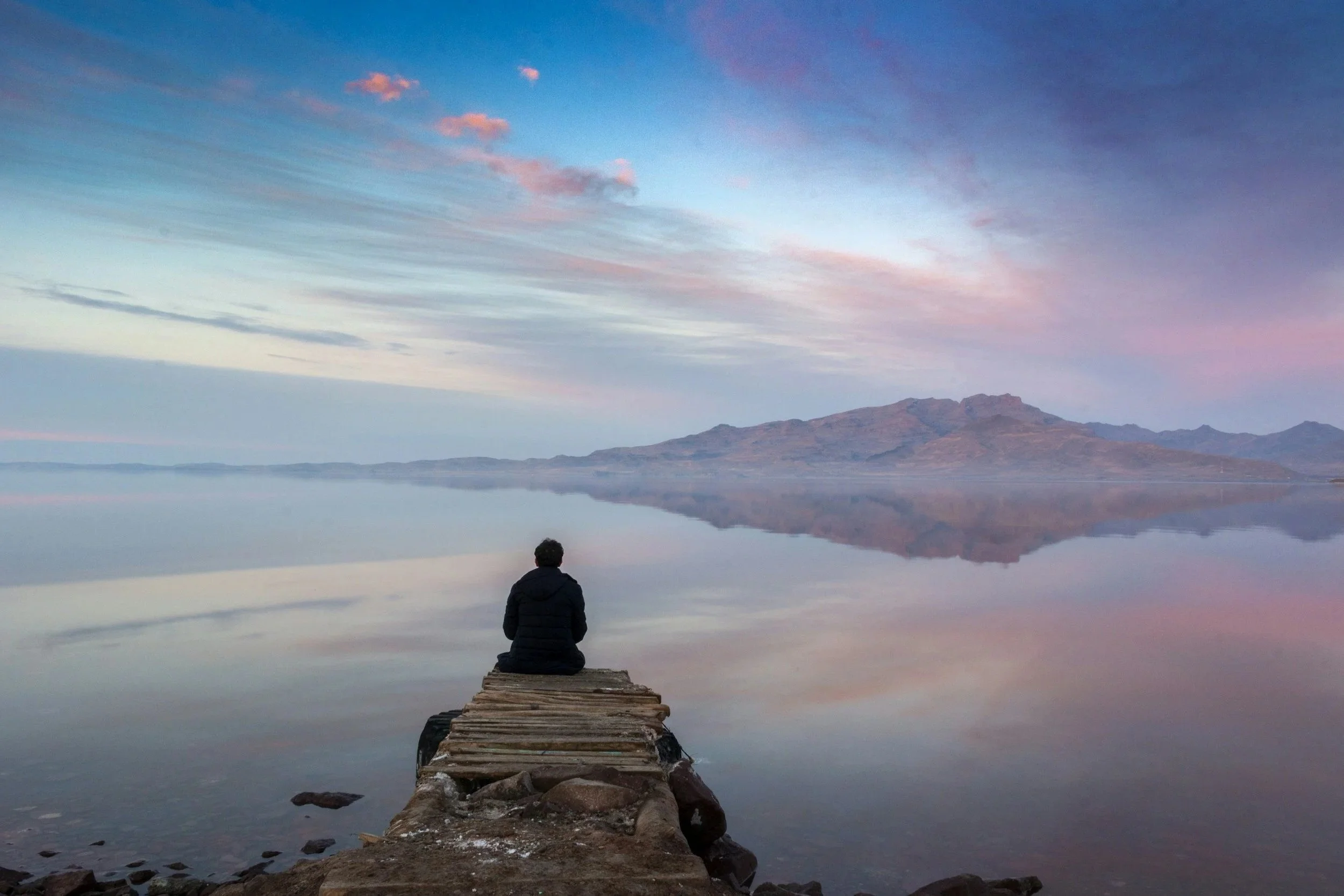 A person sitting on a wooden pier looking at a calm lake with a mountain in the background under a partly cloudy sky at sunset.