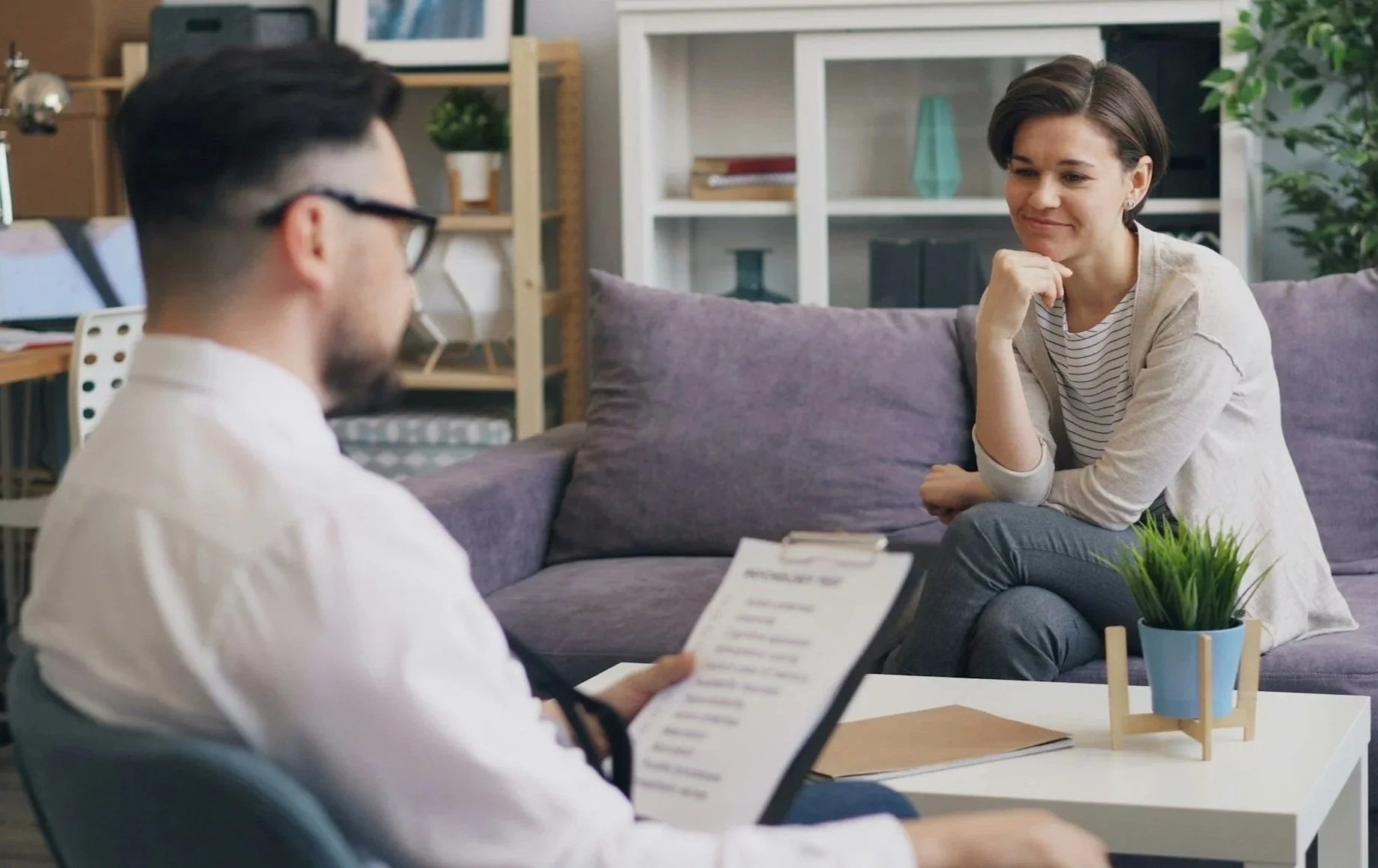 A woman in a light-colored sweater sitting on a purple couch, smiling, with her chin resting on her hand, talking to a man wearing glasses reading a clipboard, in a cozy modern living room with bookshelves and decorative plants.