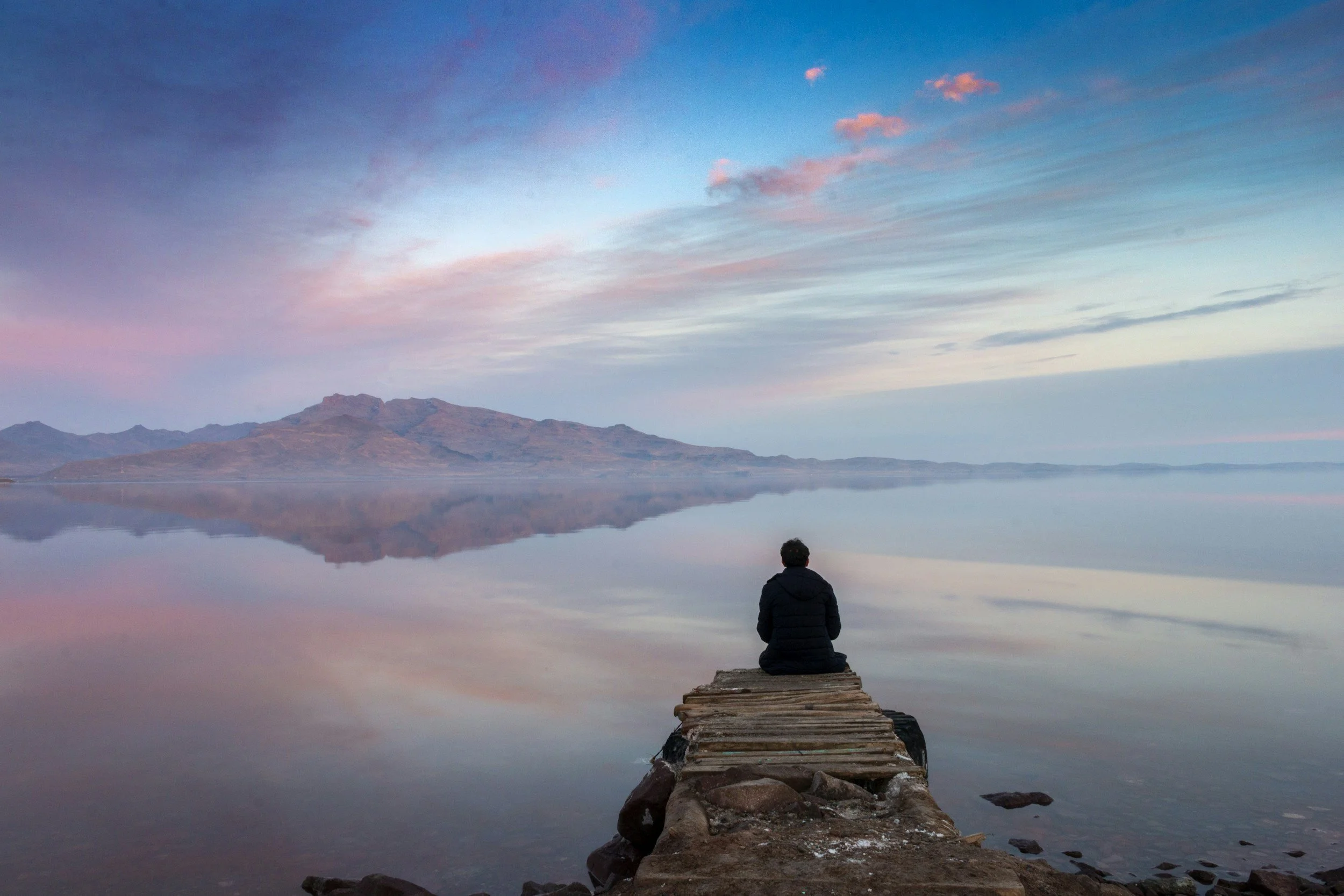 A person sitting on a wooden dock extending into a calm lake, watching a scenic sunrise or sunset with pink and purple clouds reflecting on the water and mountains in the distance.