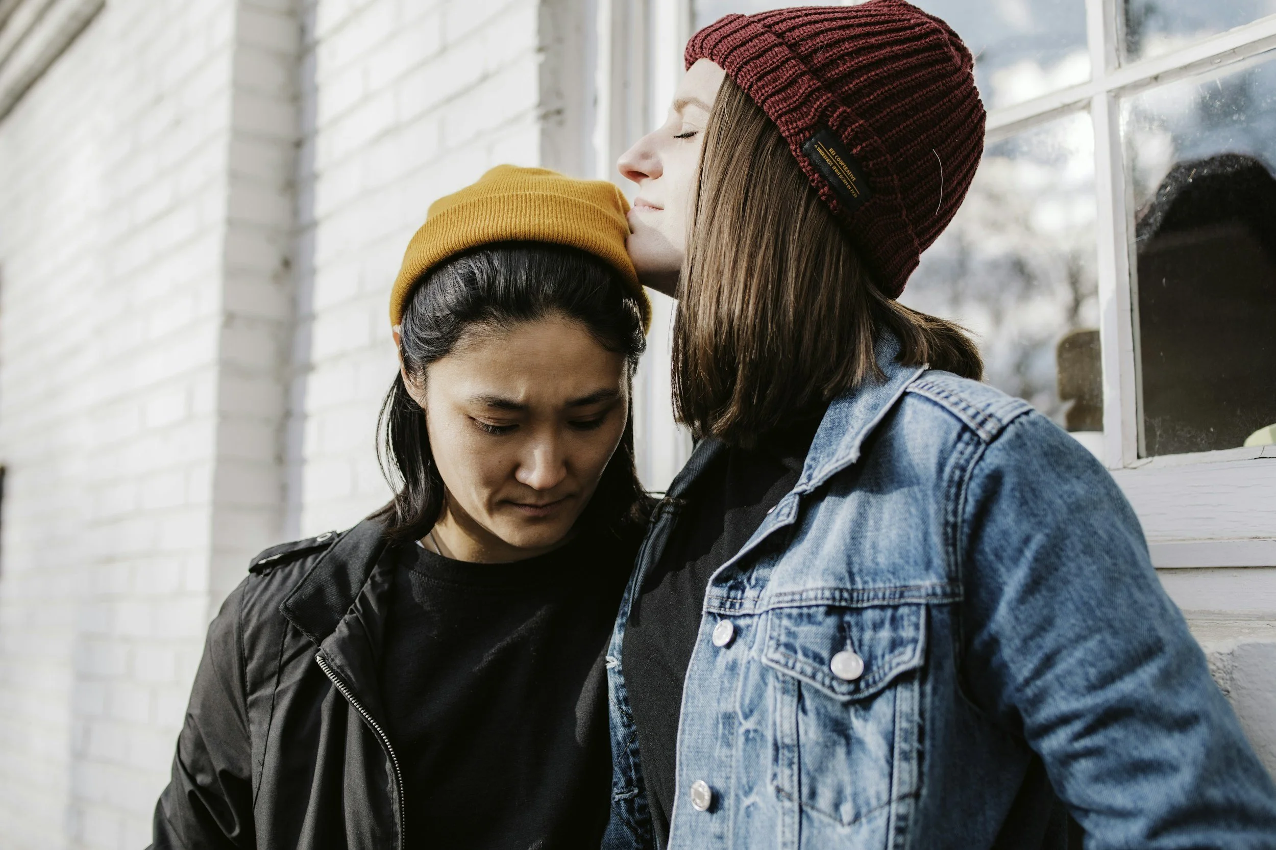 Two women wearing beanies, one red and one orange, leaning their foreheads and noses together with eyes closed in front of a white brick wall.