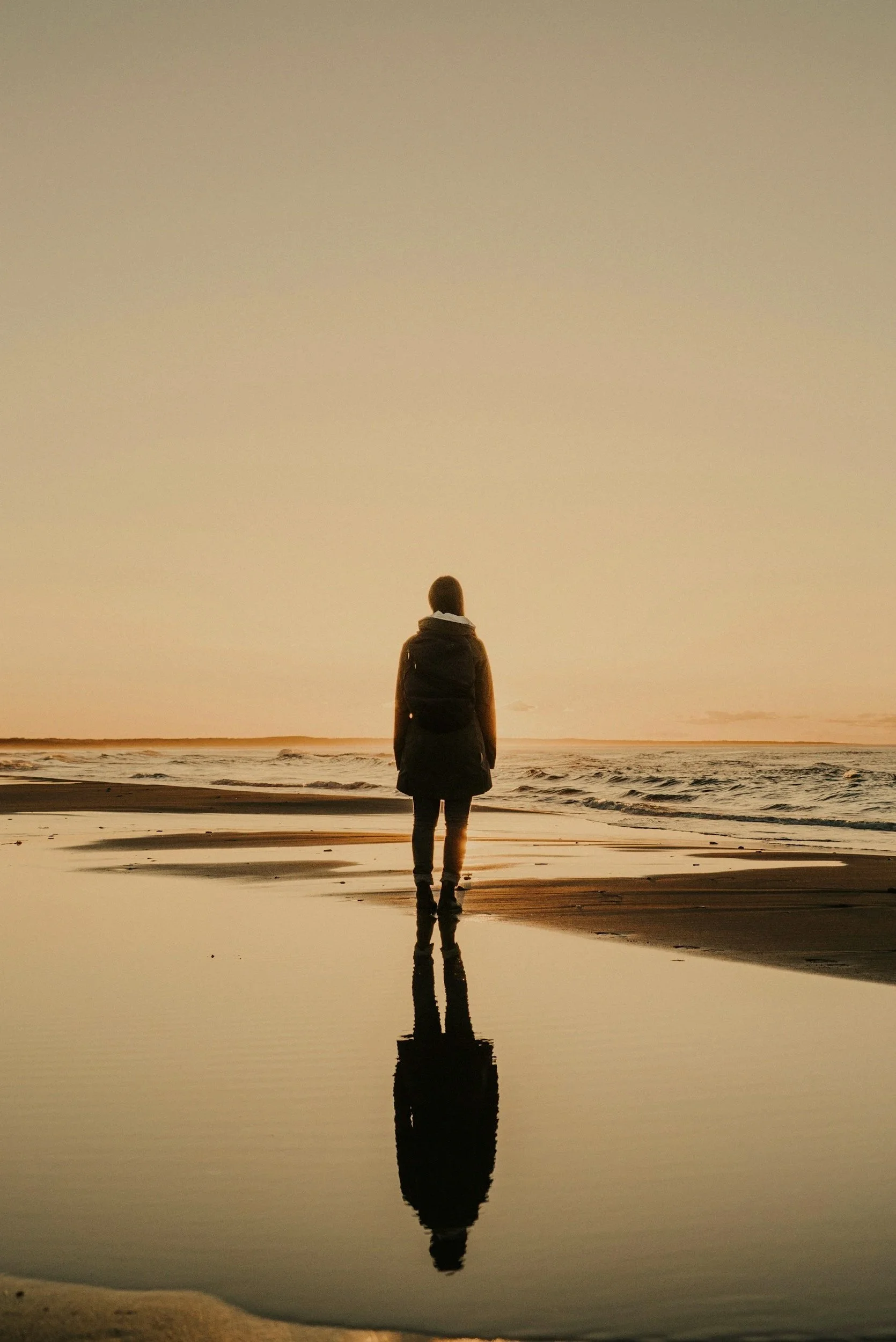 A person standing alone on a beach during sunset, with their reflection visible in a calm body of water, and the sky with a warm orange hue.