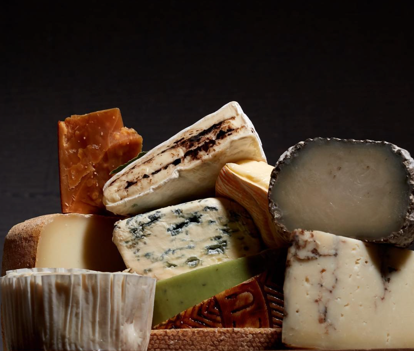 A variety of cheeses including blue cheese, brie, goat cheese, and aged cheddar arranged on a dark background.