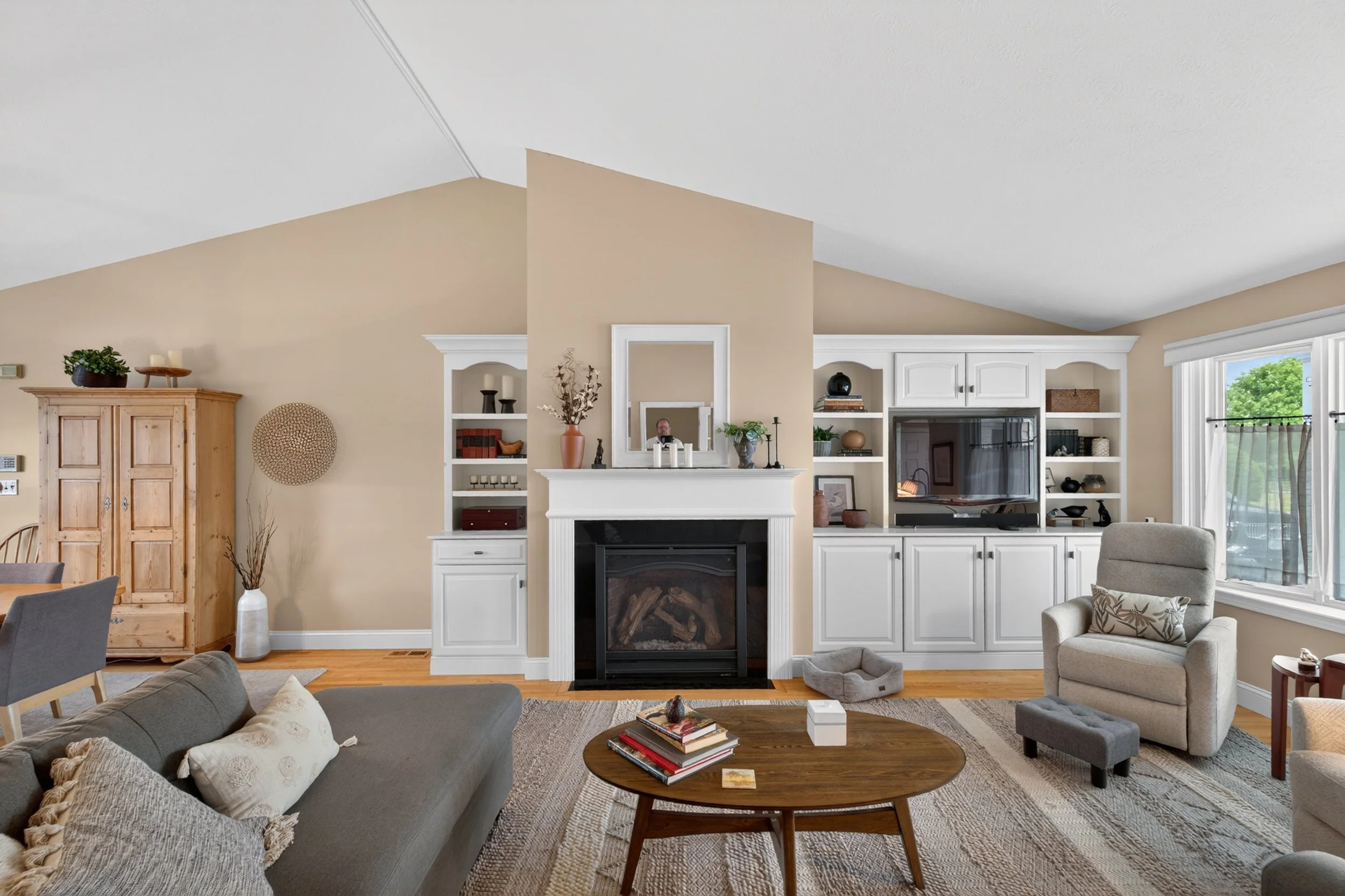 A spacious living room with a beige wall and a white fireplace, surrounded by white built-in shelves. The room has a large window with blinds and a view of green trees outside. There is a gray sofa with pillows, a wooden coffee table with books, and 