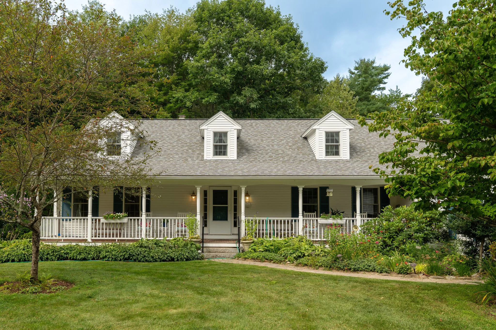 Front view of a two-story house with a gray shingled roof, white siding, and three dormer windows in a lush green yard with trees and flower garden.