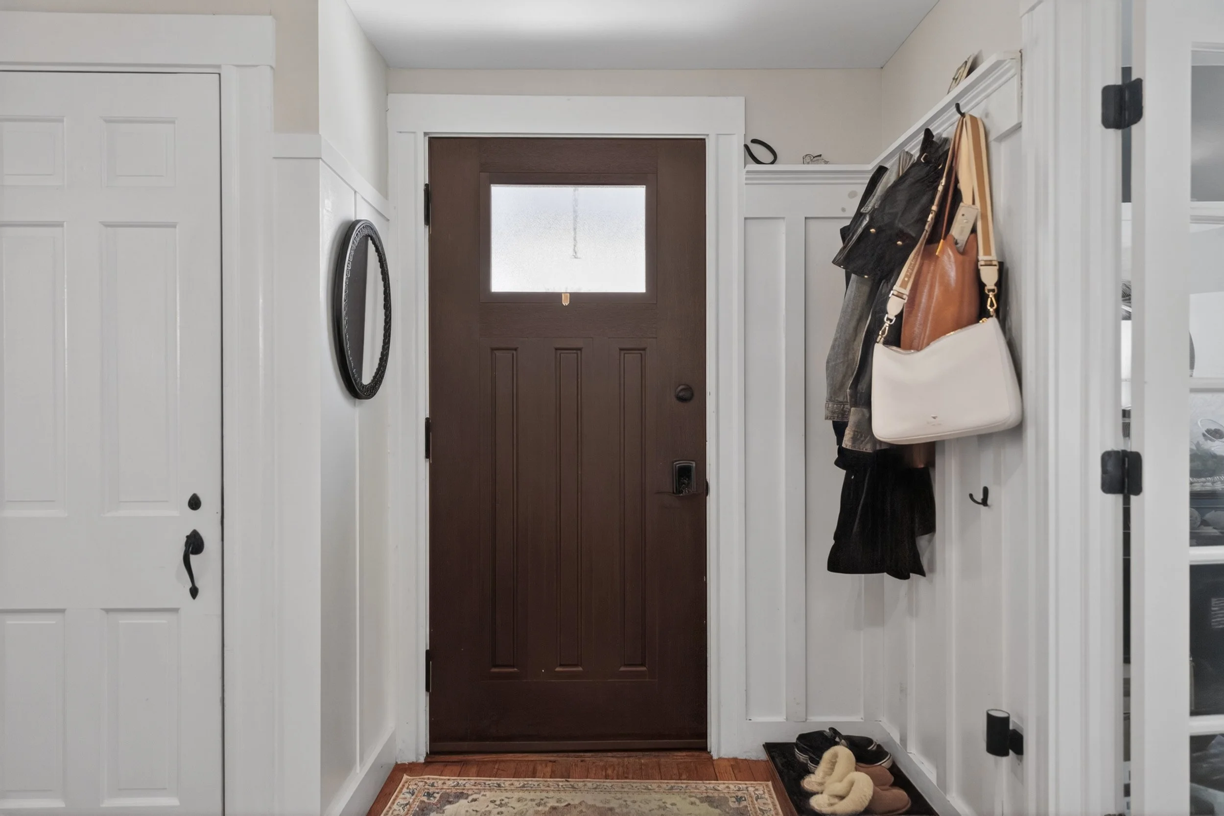 An entryway with a brown front door, white walls with paneling, coats and bags hanging on hooks on the right, a mirror on the left wall, a small rug on the hardwood floor, and shoes and slippers on the floor near the door.