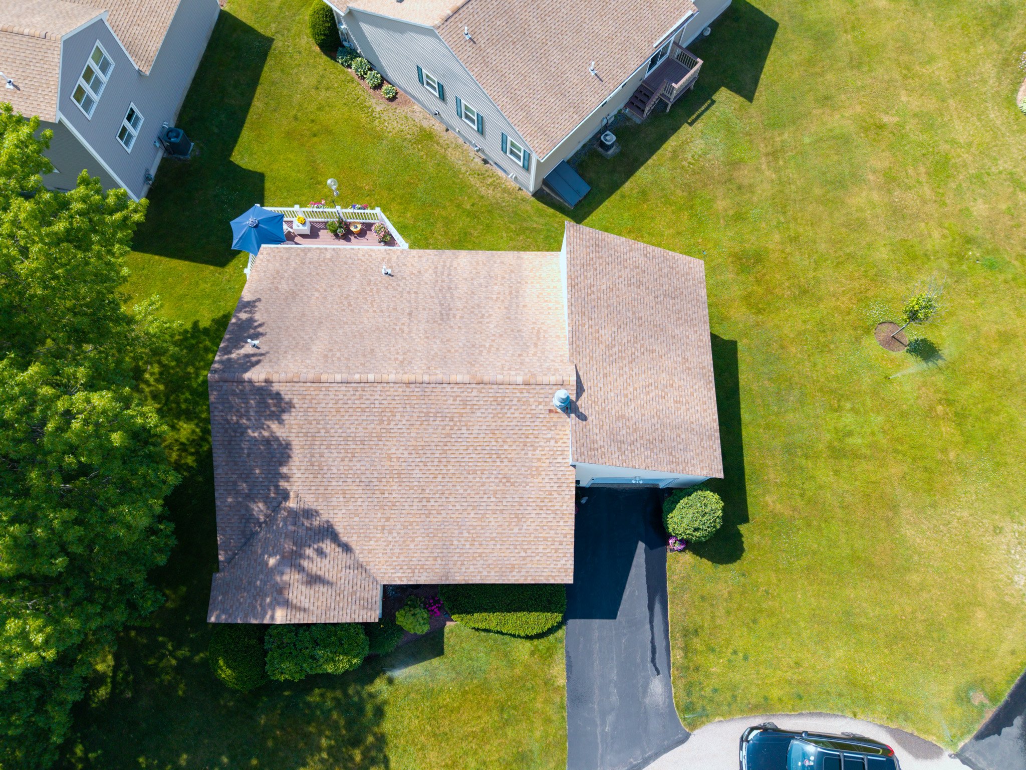 An aerial view of a residential backyard with a house featuring a brown shingle roof, a deck with outdoor furniture and an umbrella, a green lawn, and some trees.