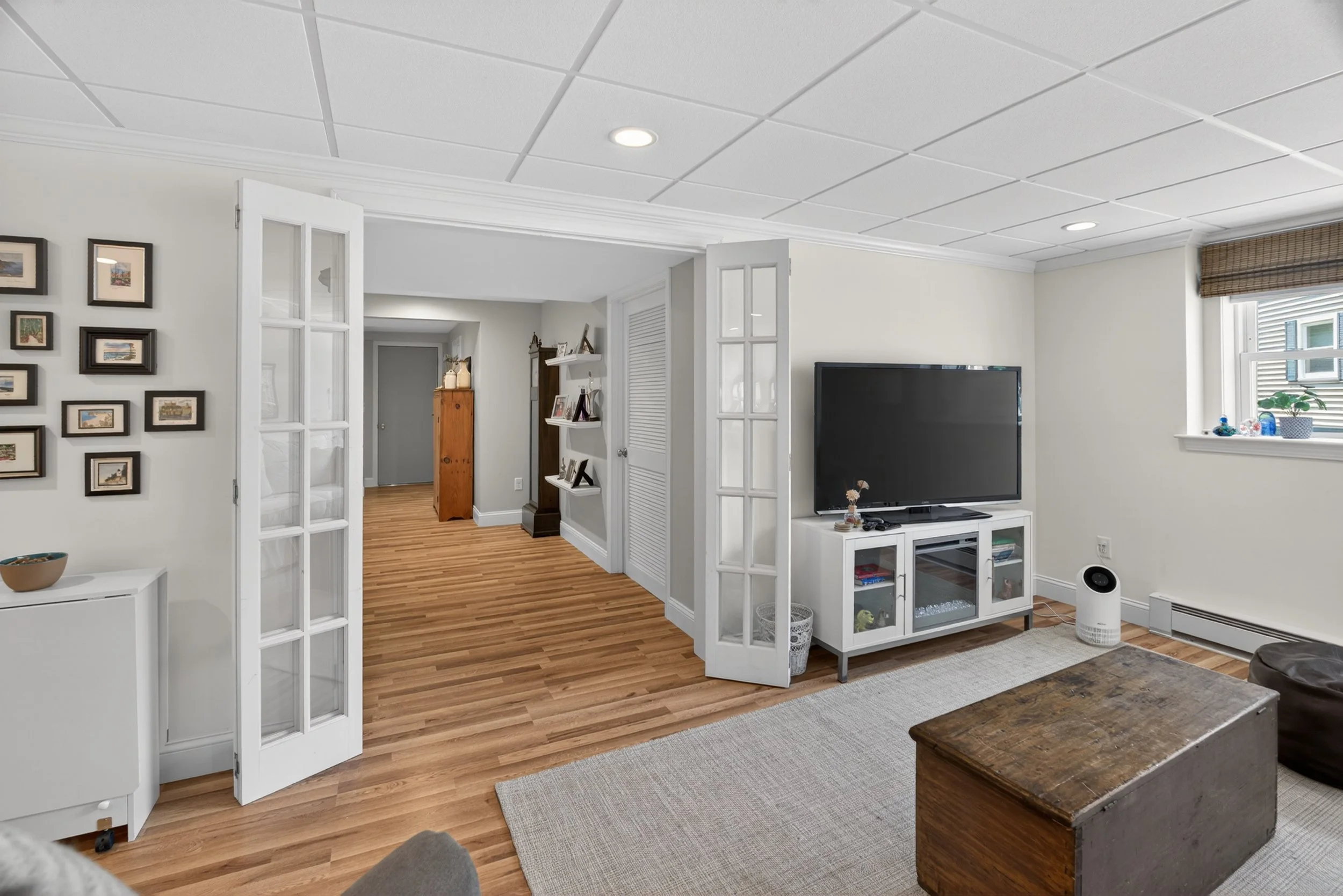 Living room with white walls, wooden flooring, and a flat-screen TV on a white stand. There's a window with a brown woven shade, framed photos on the wall, and a white folding screen door leading to another part of the house.