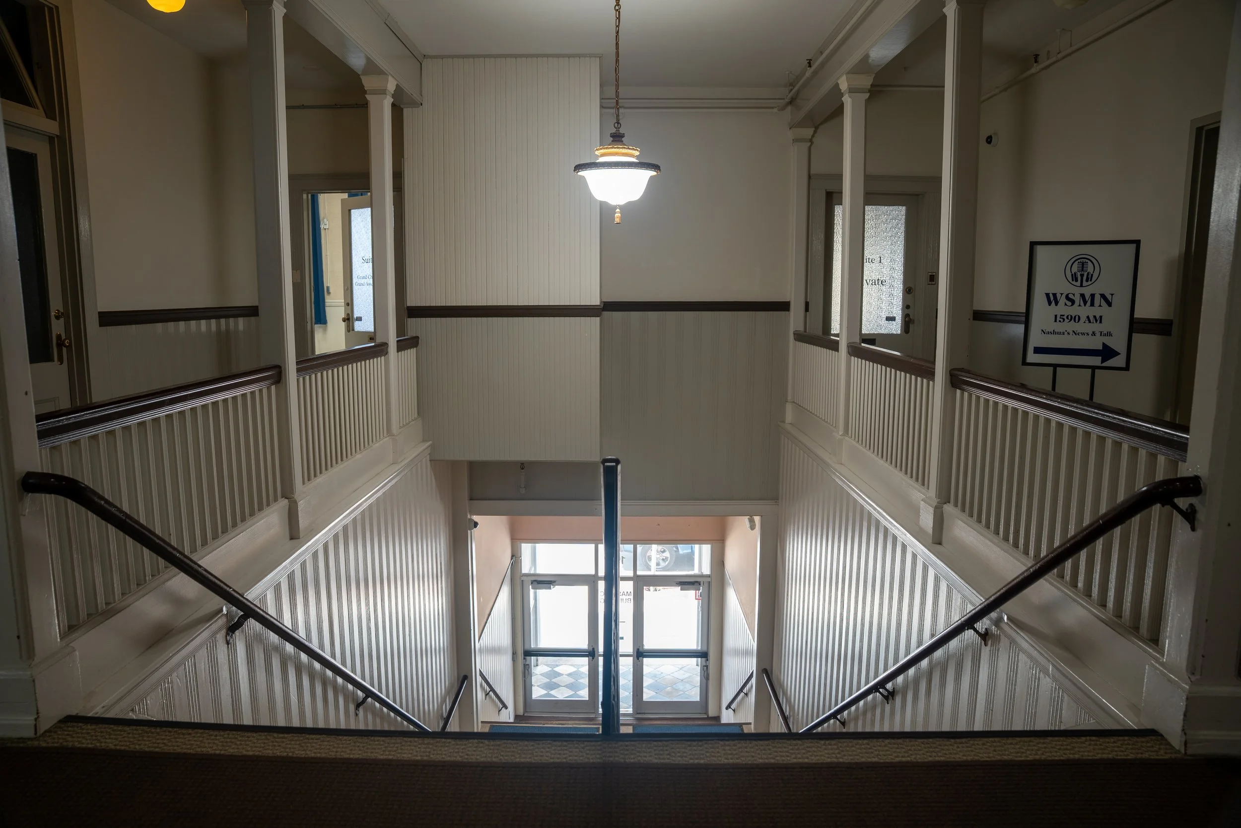 Interior view of a staircase and hallway area in a building, with a hanging light fixture, railings, and doors leading outside.