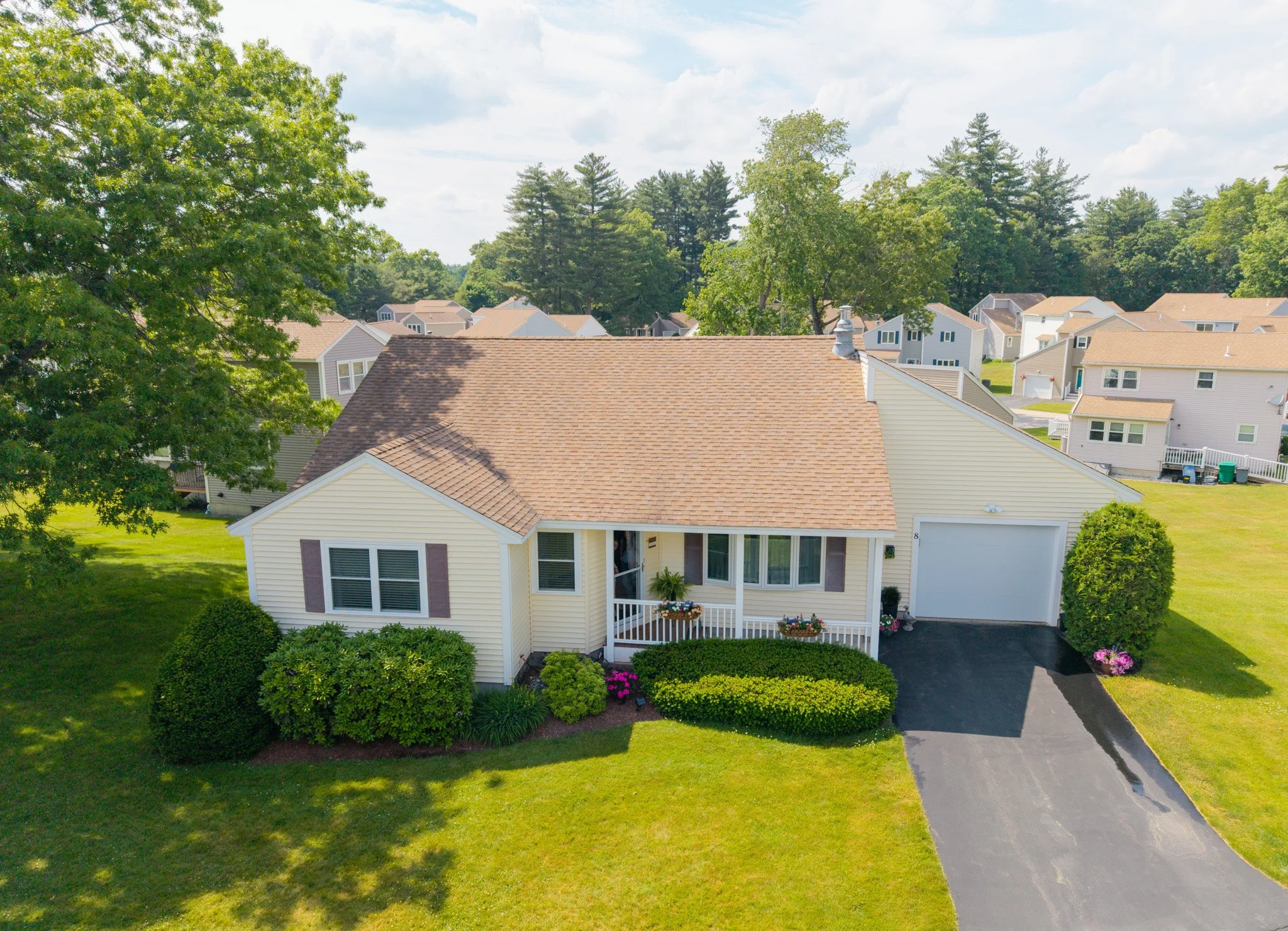An aerial view of a two-story house with a yellow exterior, brown shingle roof, front porch with flower pots, surrounded by green lawns, bushes, and trees, in a neighborhood with other houses and tall trees in the background under a partly cloudy sky